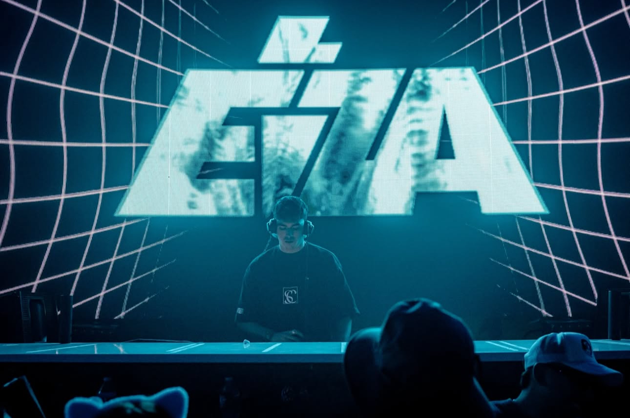 DJ performs on stage with a large illuminated logo behind him, audience in foreground, grid pattern on screen, dark atmosphere.