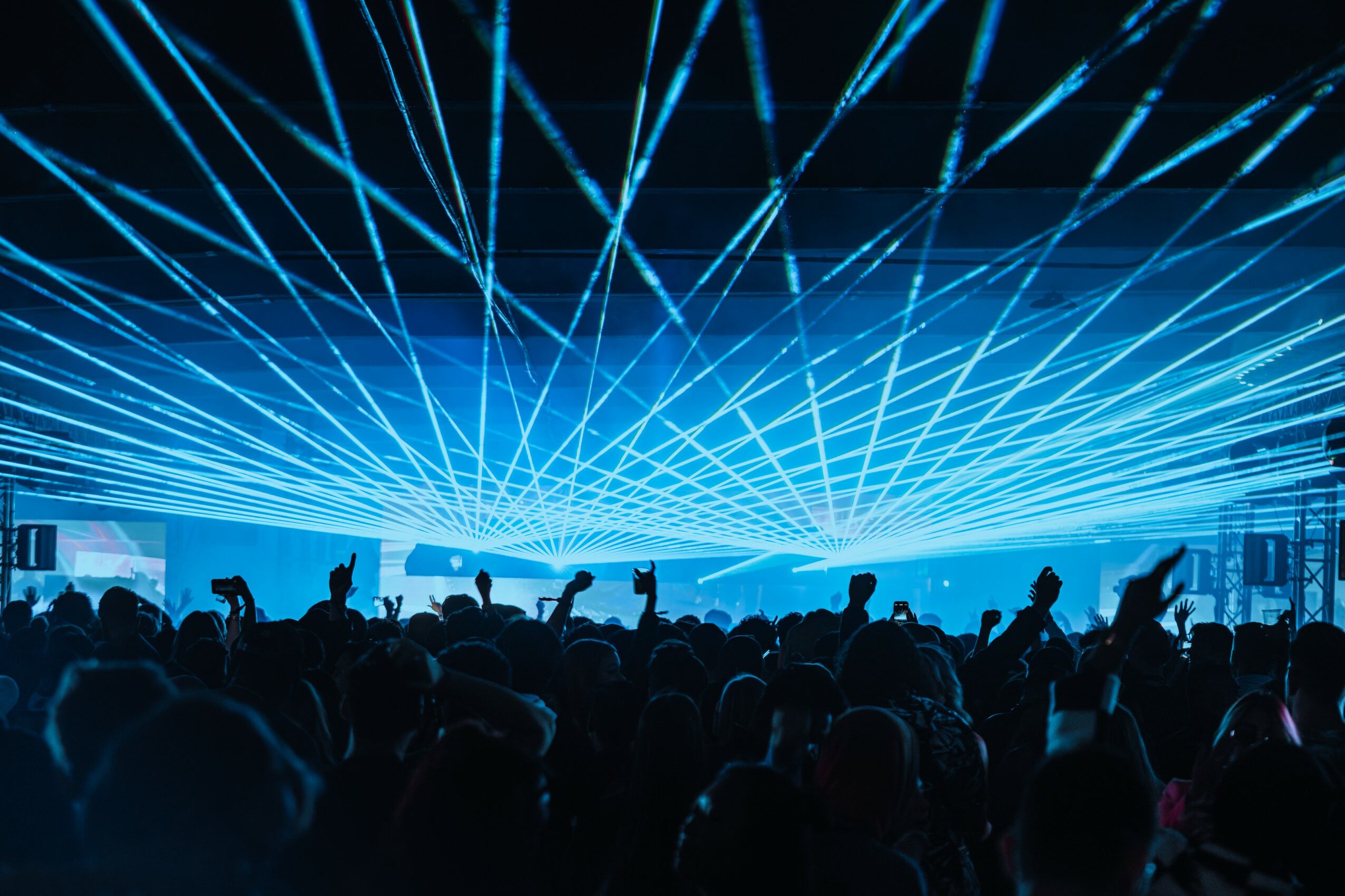 Crowd at concert with blue laser lights overhead, people raising hands, dark background, lively atmosphere.