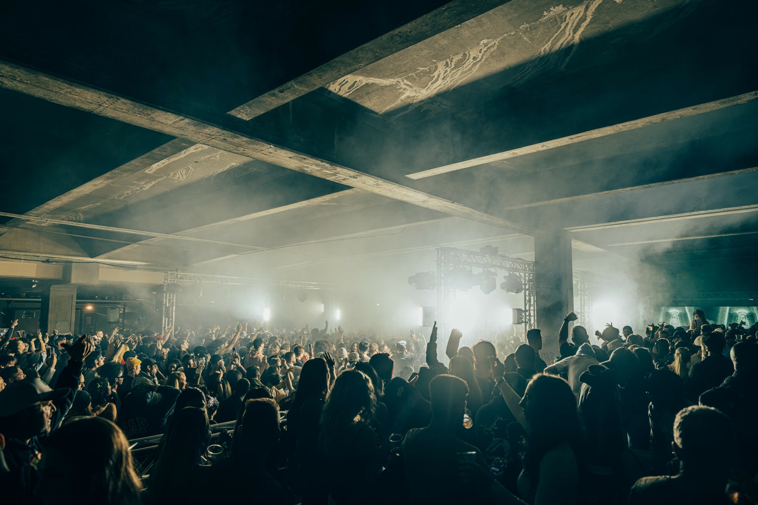 Crowd of people at a concert or event with bright stage lights and smoke effects, silhouettes visible, indoors.