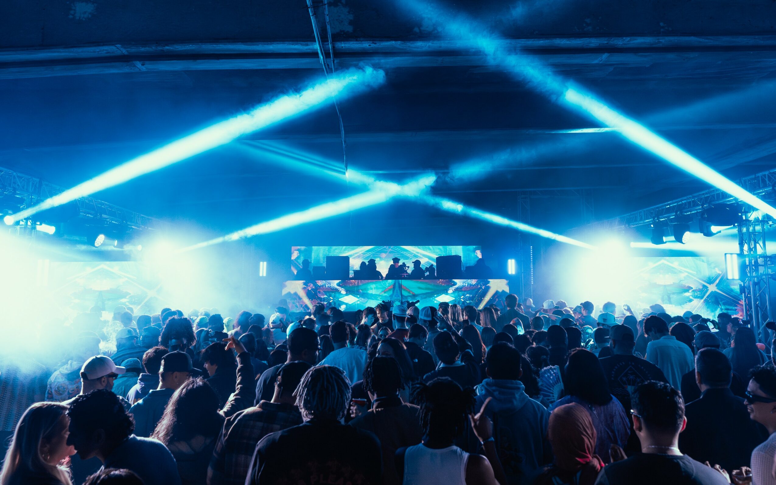 Crowd at a concert with bright blue stage lights and a large screen, people facing the stage, dark environment.