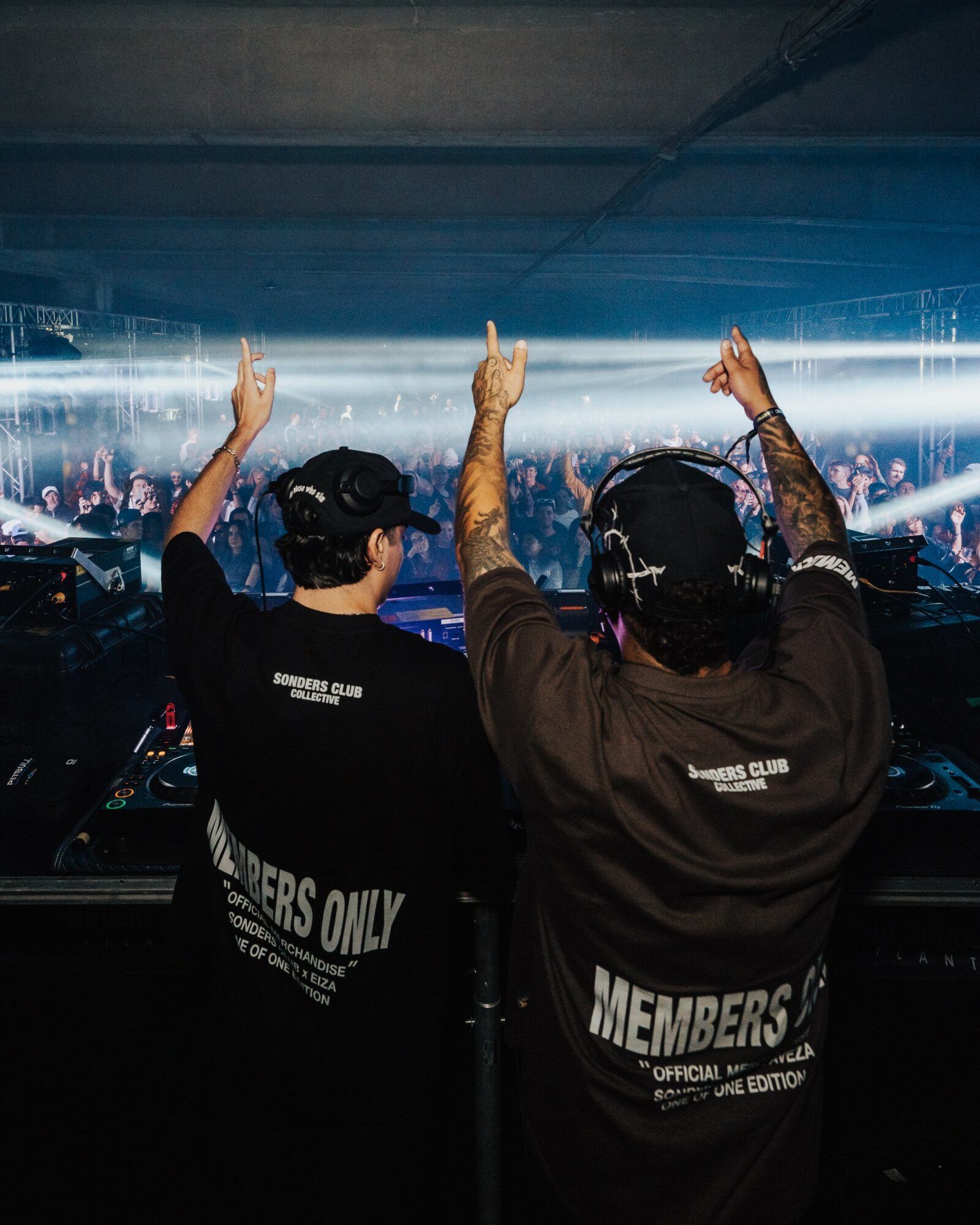 Two people in DJ booth raising hands at a concert with crowd and light effects in background.