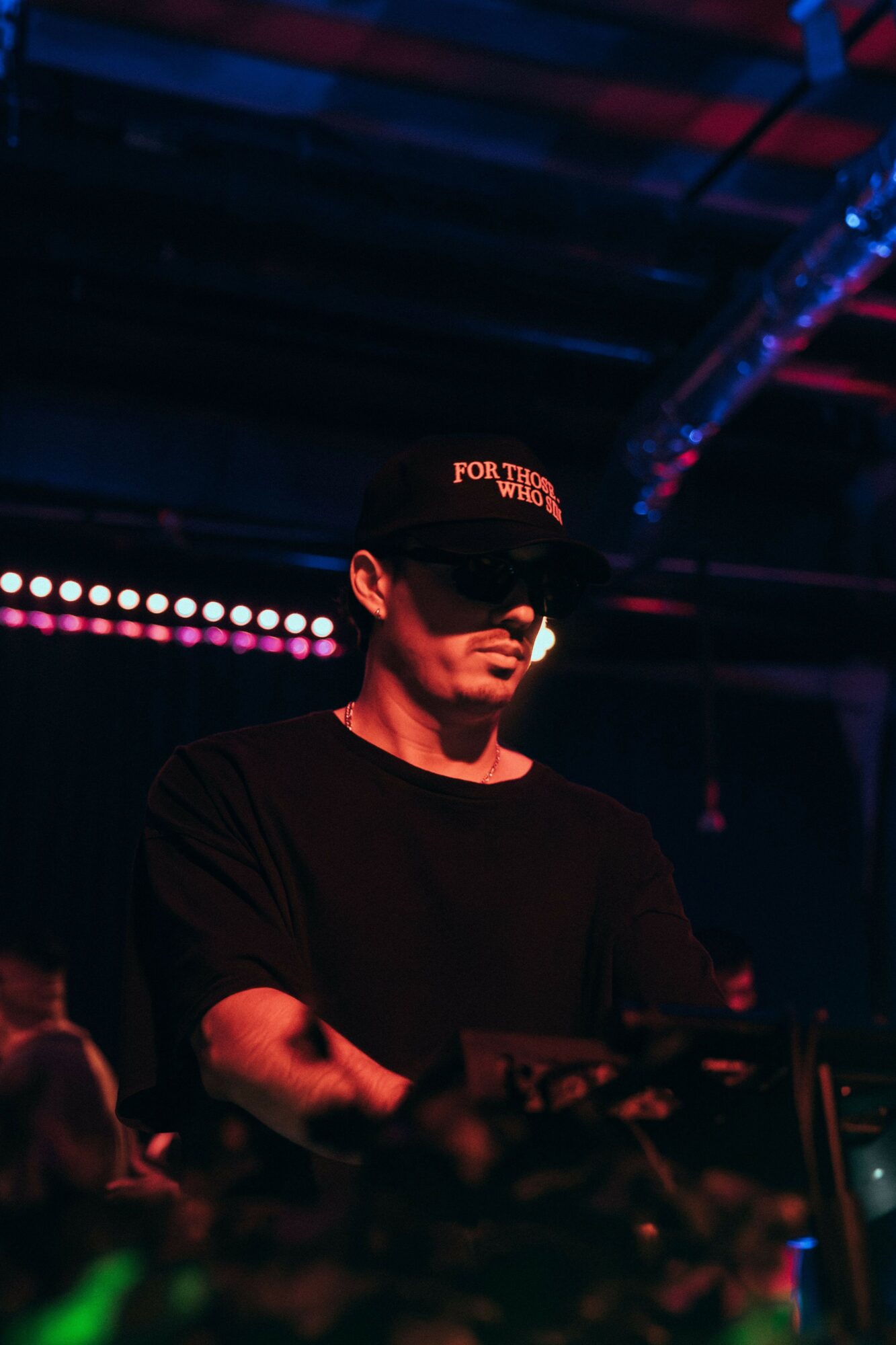 Person wearing a black cap and shirt, standing in a dark environment with colorful lights, possibly a stage or club.