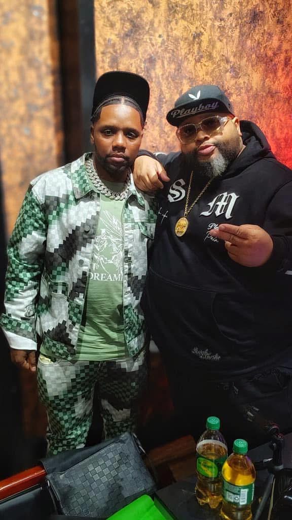 Two men pose together indoors, one wearing a patterned green and white outfit, the other in a black hoodie and cap, with drinks on a table.