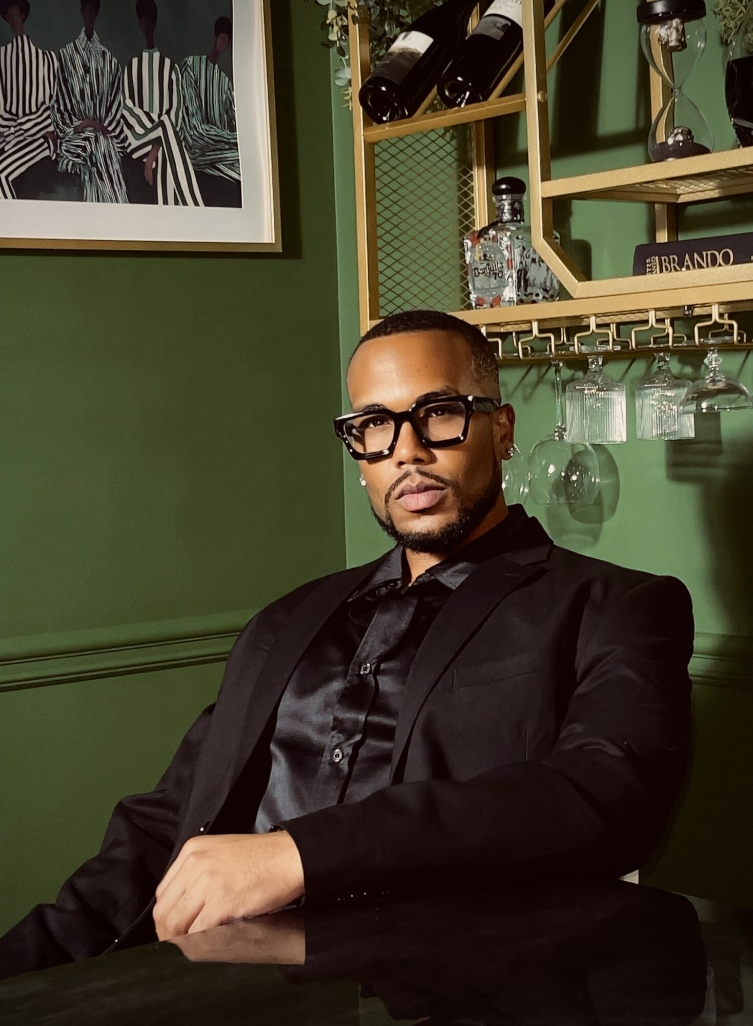 Man in black suit and glasses sitting in a room with green walls and shelves with bottles and glasses.