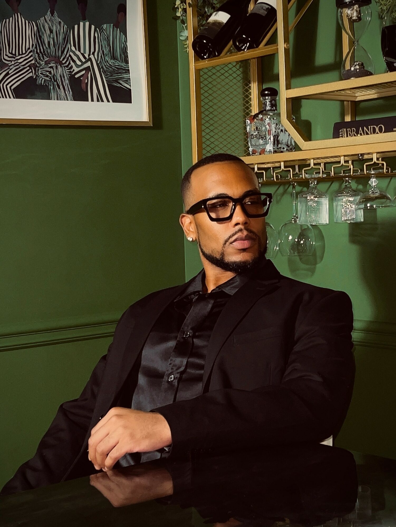 Man with sunglasses in a black suit sitting at a bar with green walls and shelves with bottles and glasses