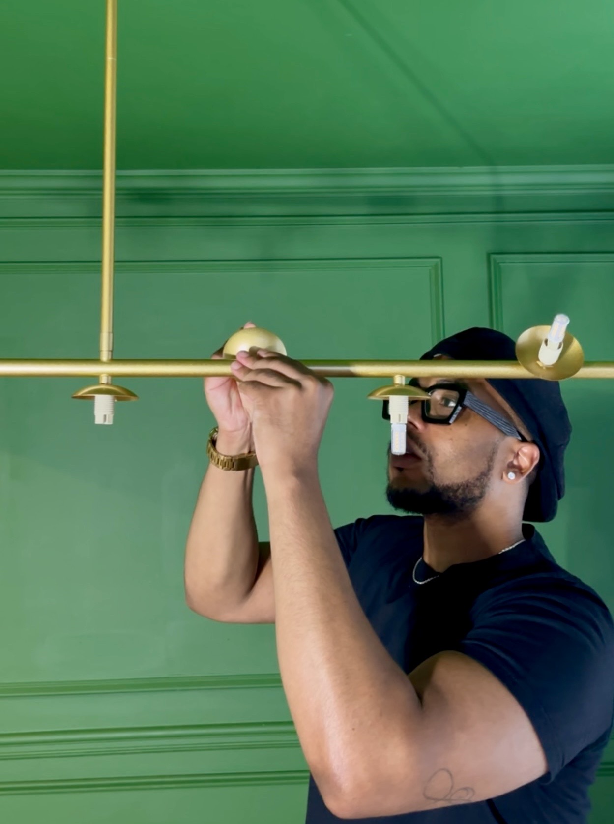 Man with glasses and a beard adjusts a gold-colored ceiling light fixture against a green wall.