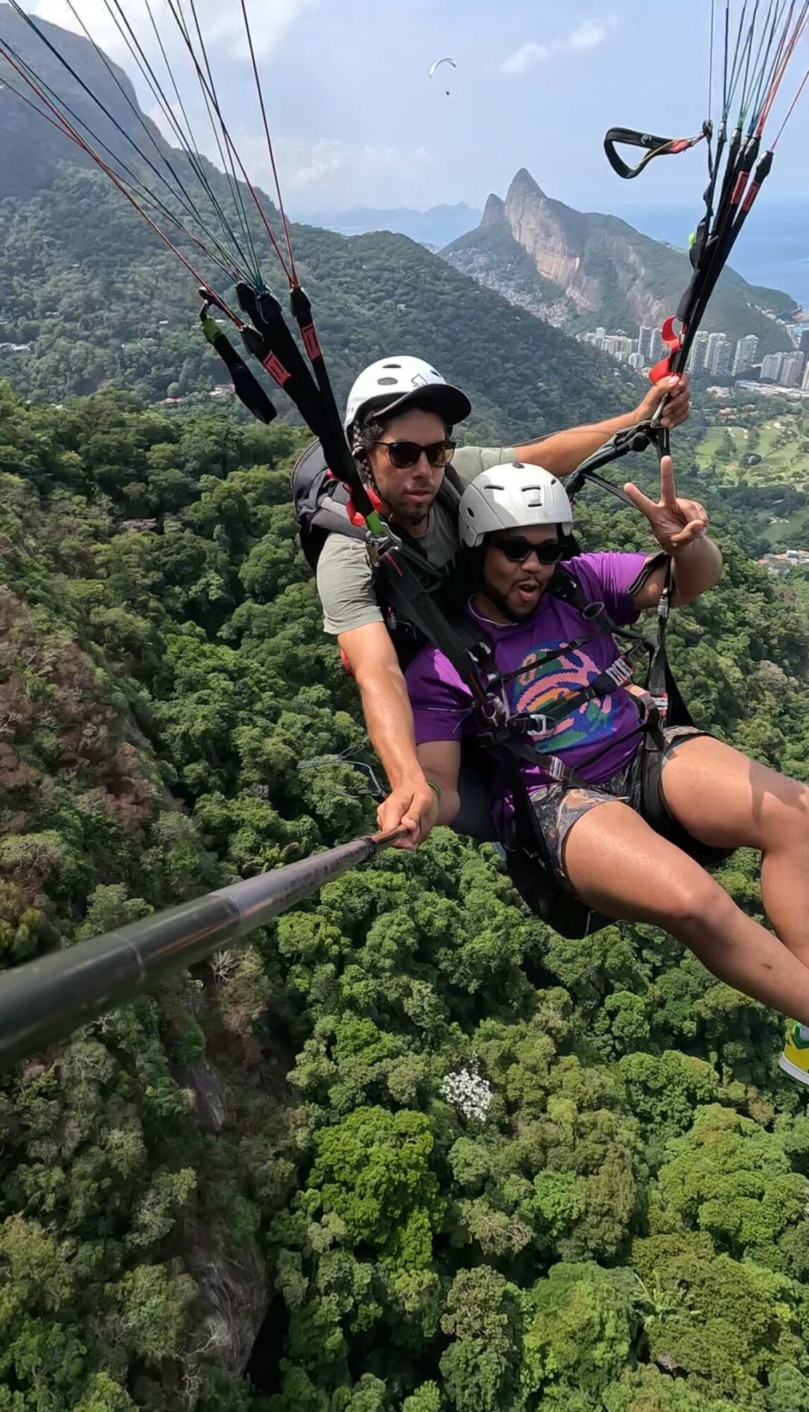 Two people paragliding over a lush green forest with mountains in the background, one making a peace sign, both wearing helmets.