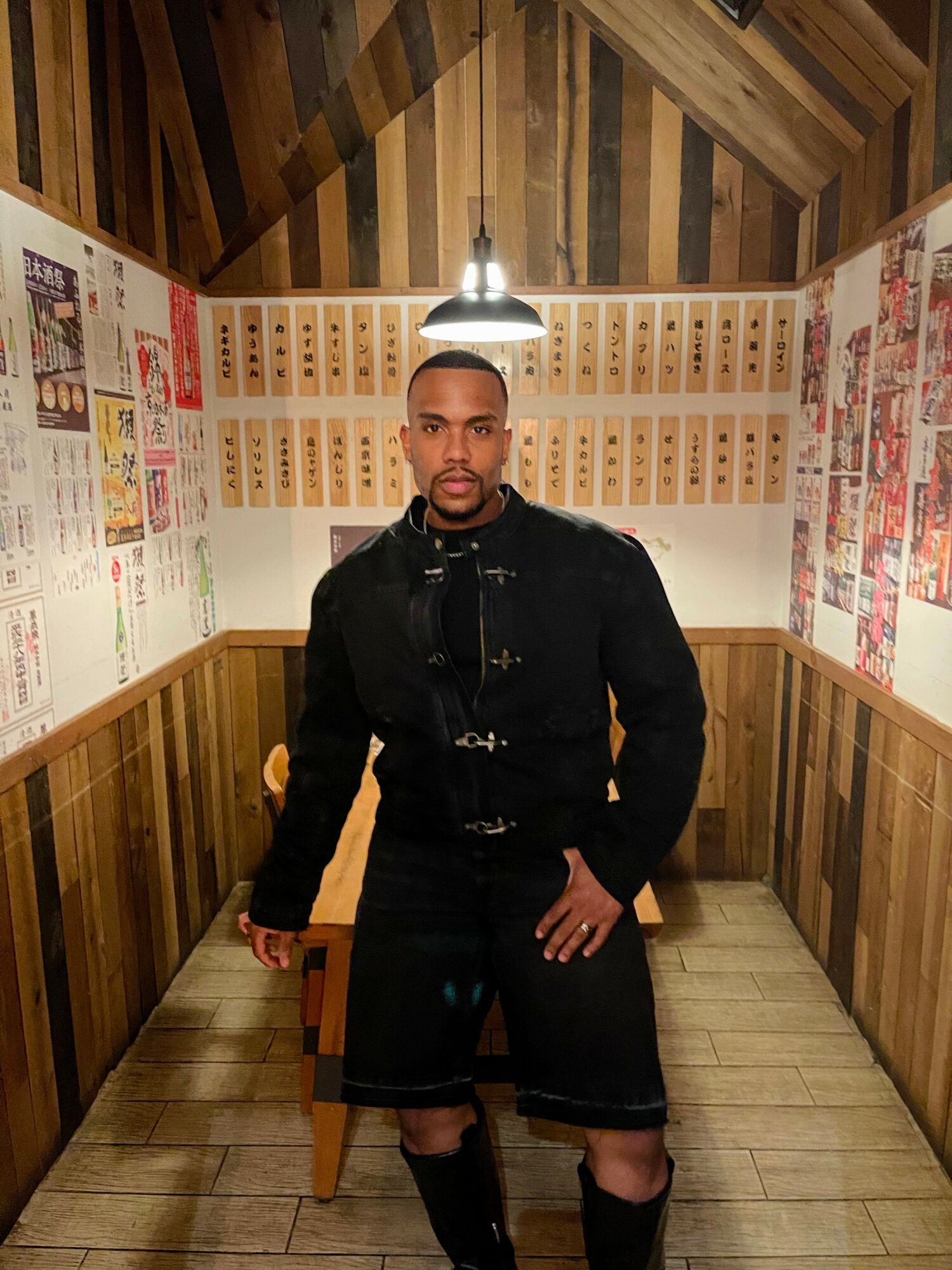 Young man standing in a wooden-paneled room with posters on the walls, wearing a black jacket and shorts.