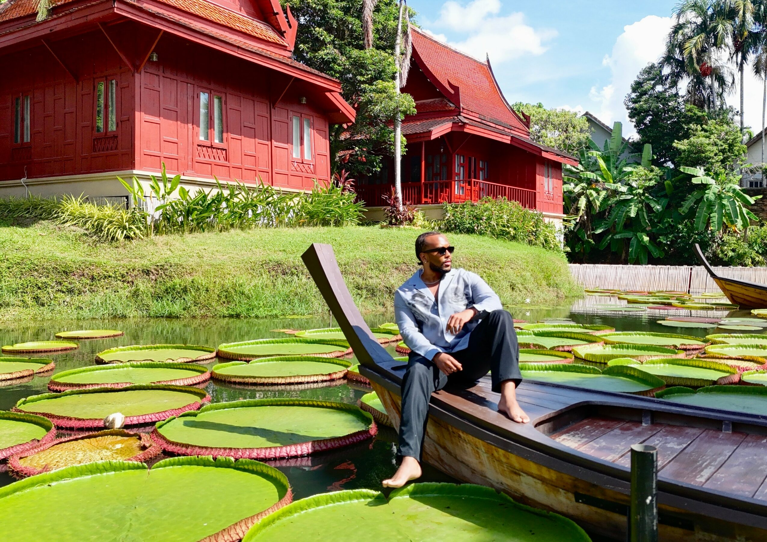 Man sitting on a boat surrounded by large green lily pads, with traditional red buildings and trees in the background.
