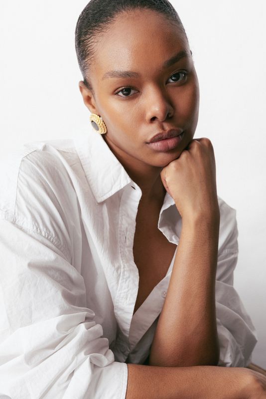 Young woman with dark hair in a bun, wearing a white shirt and gold earrings, resting her chin on her hand.