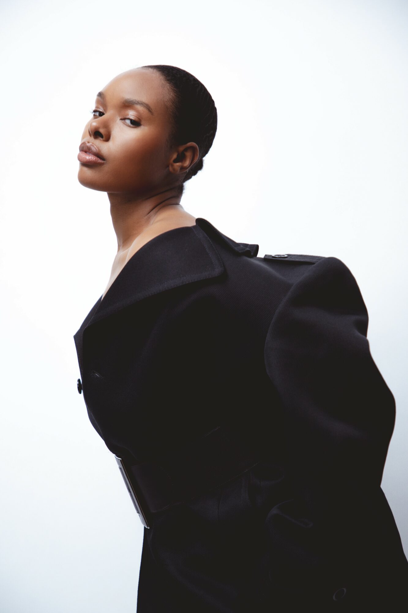 Portrait of a woman with short hair wearing a black coat, looking over her shoulder against a white background.