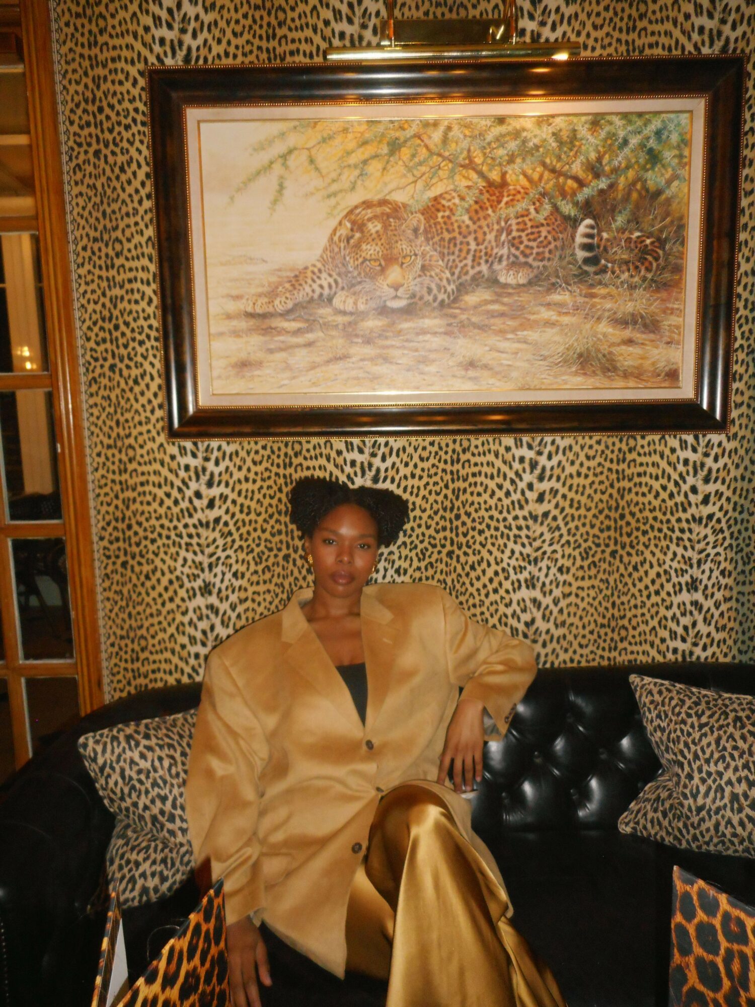 Person sitting on a black chair in a room with leopard print walls and a large framed painting of a leopard, wearing a gold outfit.