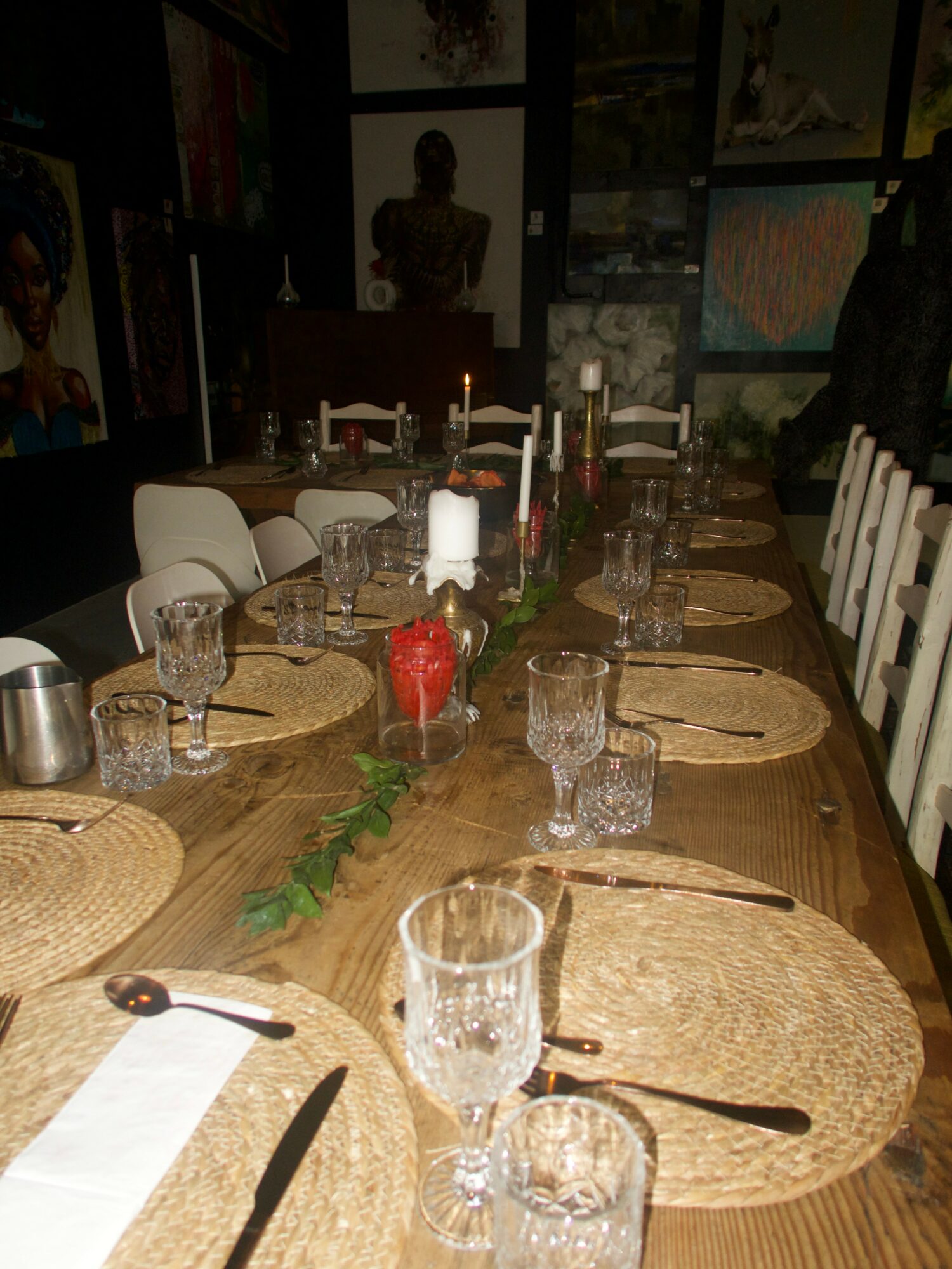 Long dining table set with placemats, glasses, cutlery, and a centerpiece with candles and greenery, in a dimly lit room.