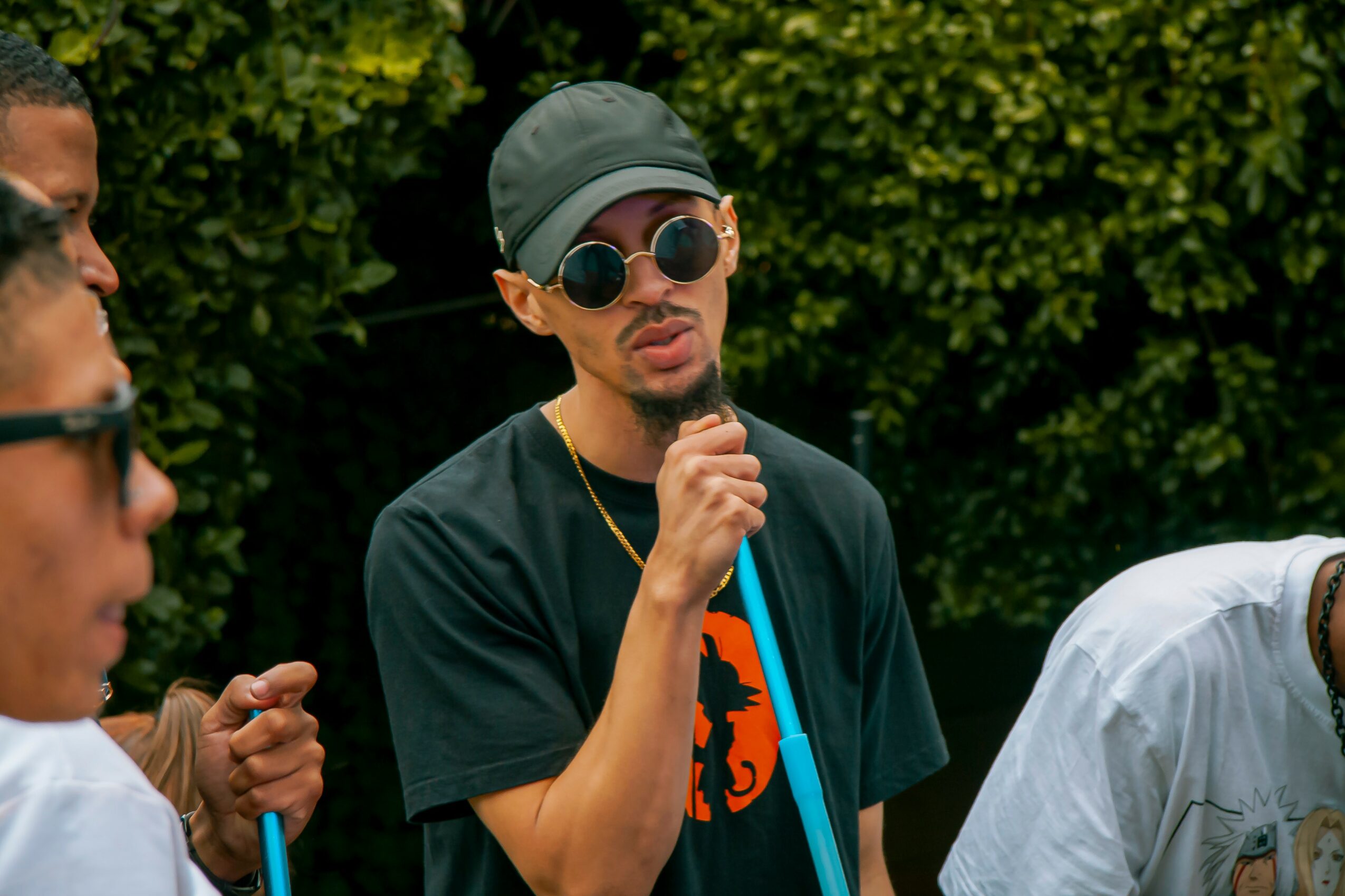 Young man wearing sunglasses, a cap, and a black T-shirt with an orange graphic, holding a microphone outdoors with greenery background.