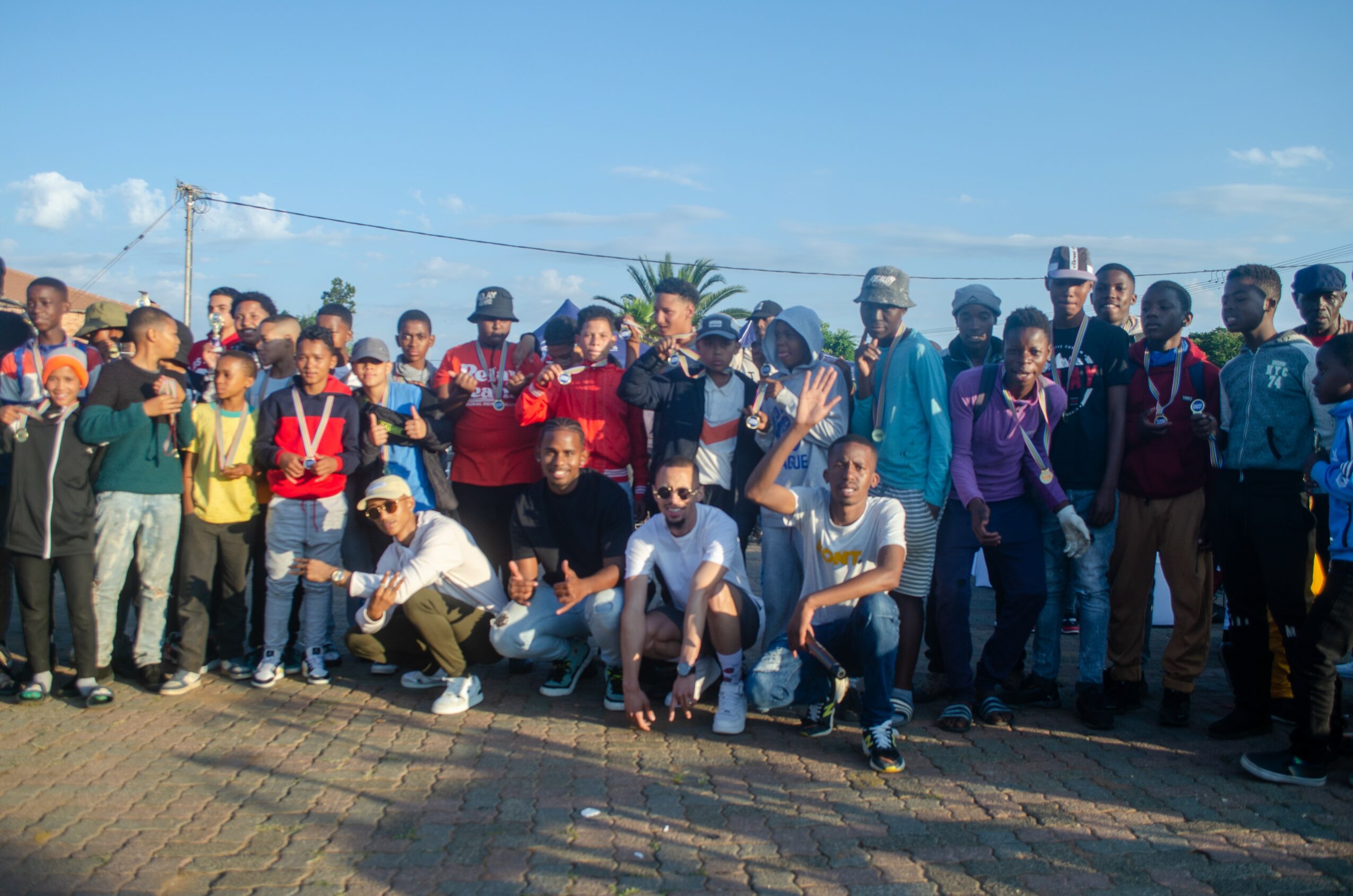 Group of people outdoors, some wearing medals, standing and sitting on a paved area, with a blue sky and trees in background.