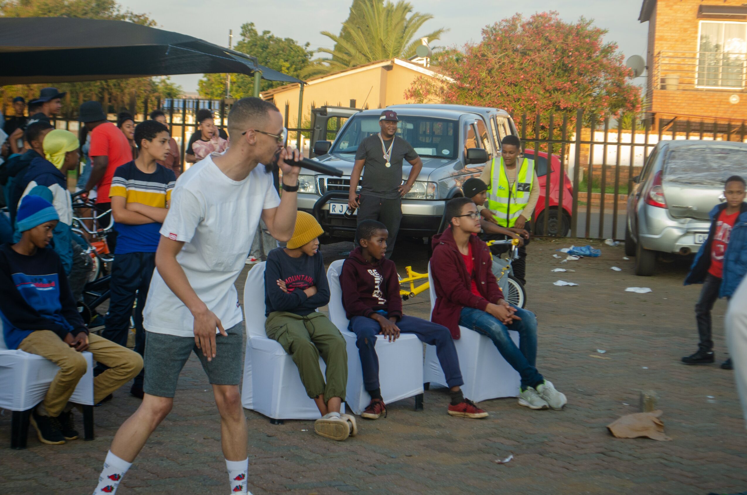 Group of people sitting and standing outdoors, some talking, with cars and a building in the background.