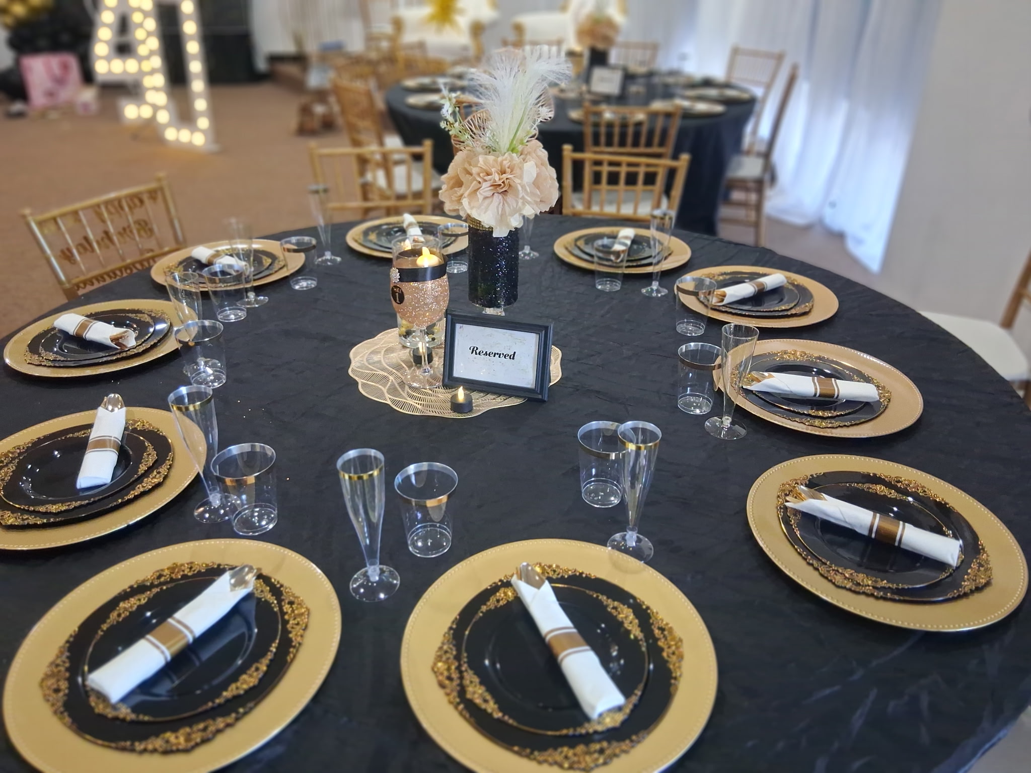 Round table set with black and gold plates, glasses, and a floral centerpiece, in a decorated event space.