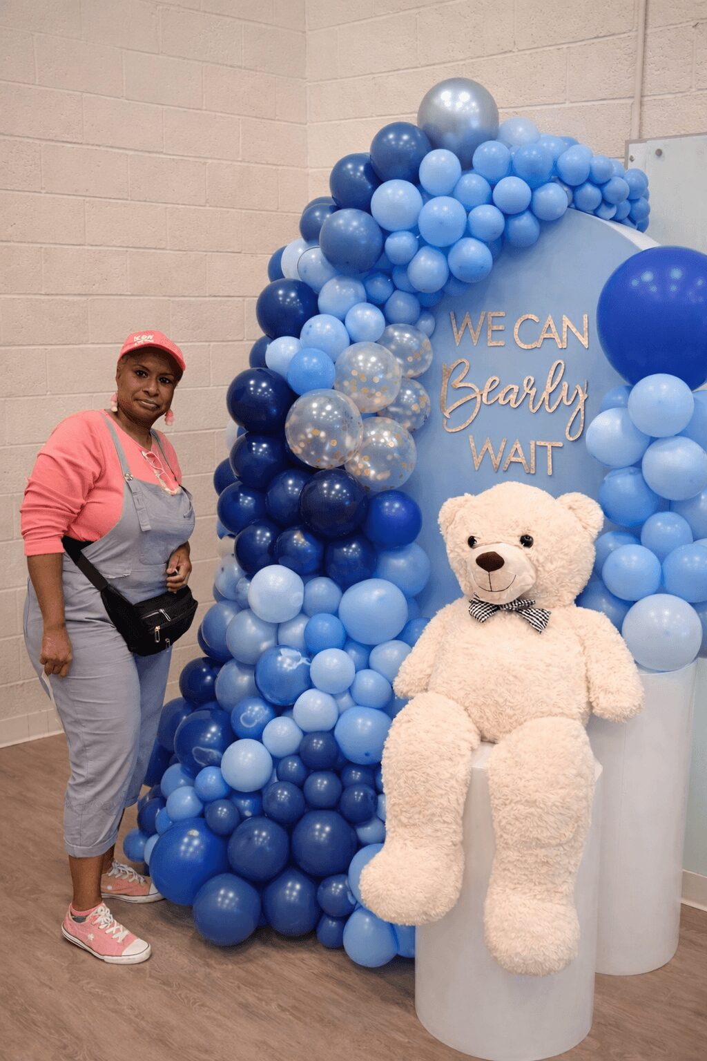 Woman standing next to a balloon arch and a large teddy bear, with a sign reading 'We can clearly wait'.