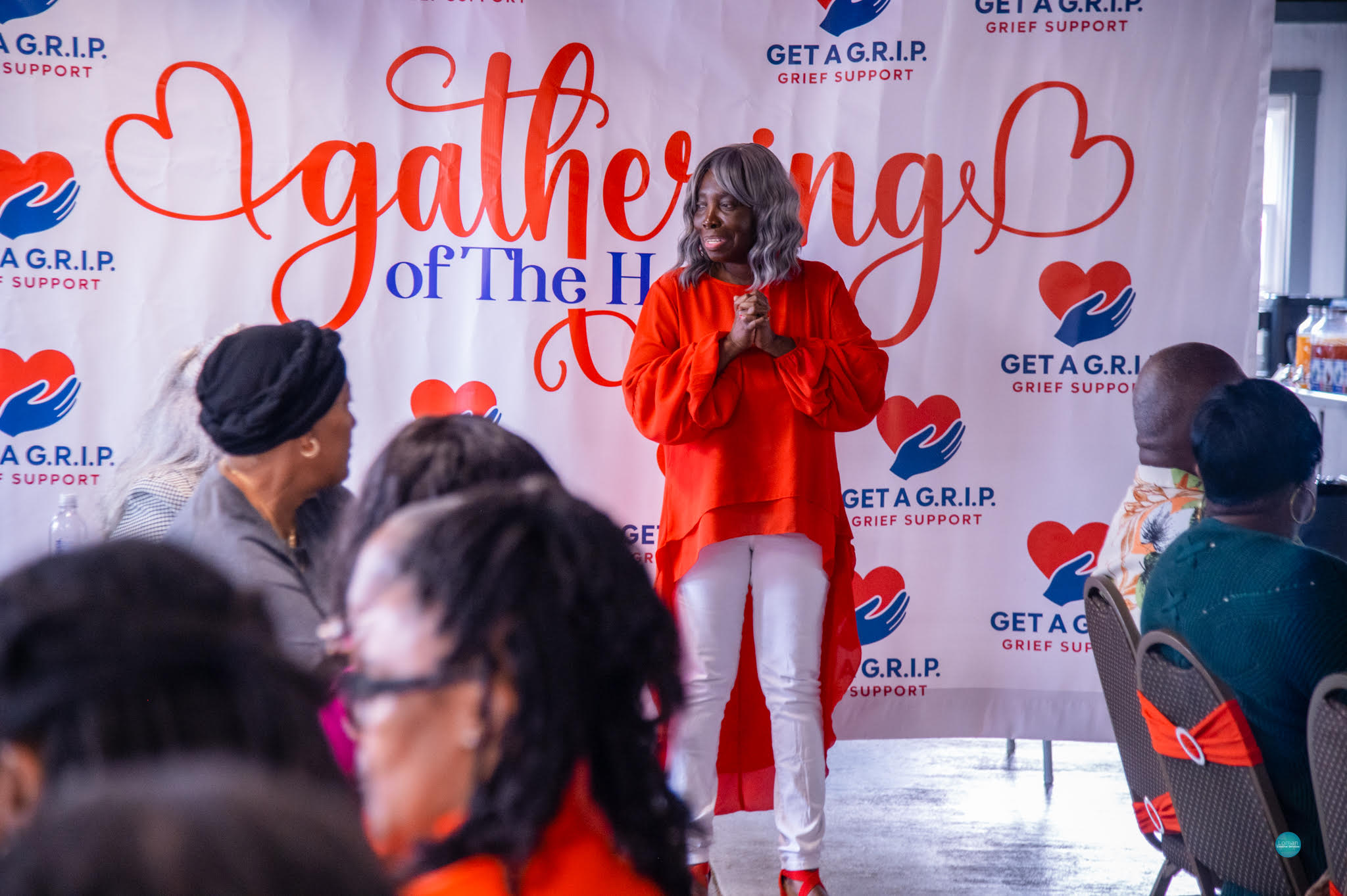 Woman speaking on stage at event with audience seated, backdrop reads 'Gathering of The Heart' with hearts and logos.