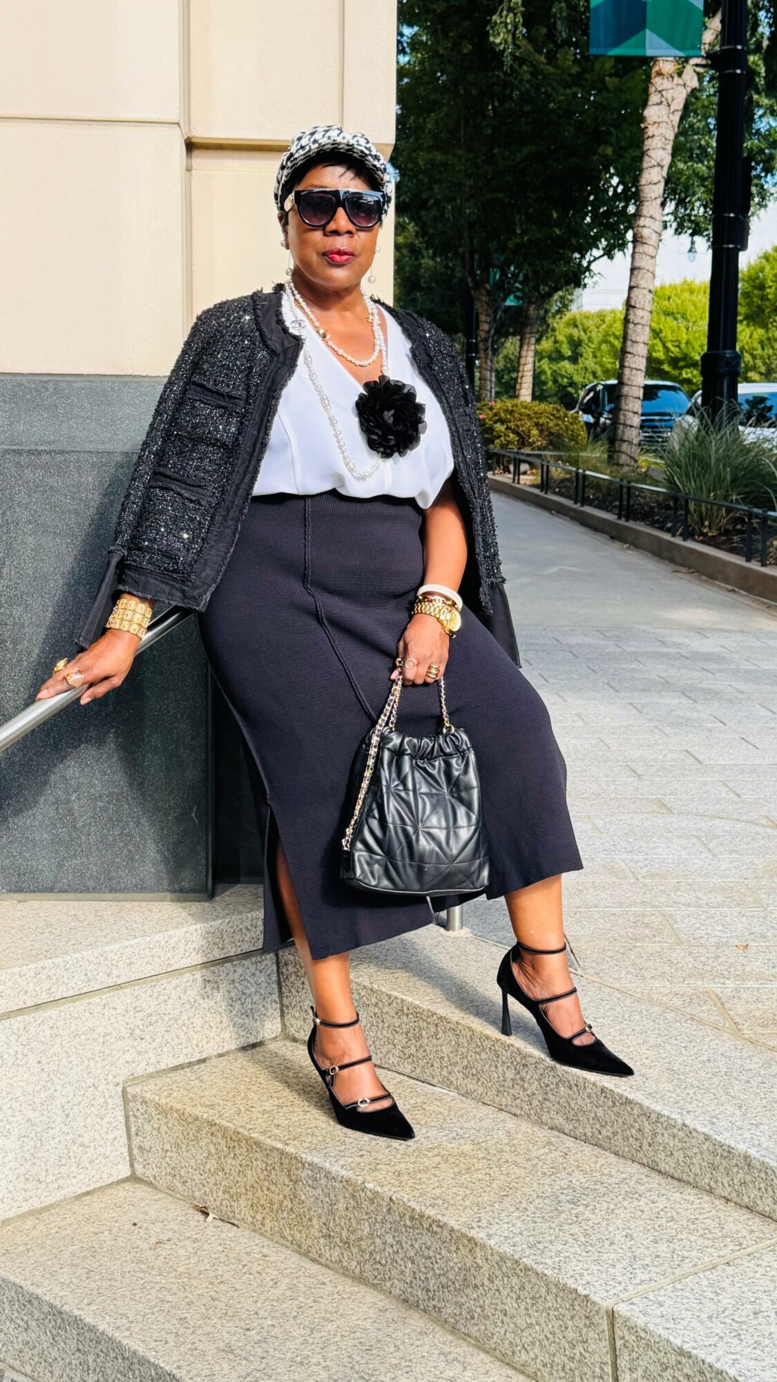 Woman sitting outdoors on steps, wearing sunglasses, a black blazer, white blouse, dark pants, and high heels, holding a black handbag.