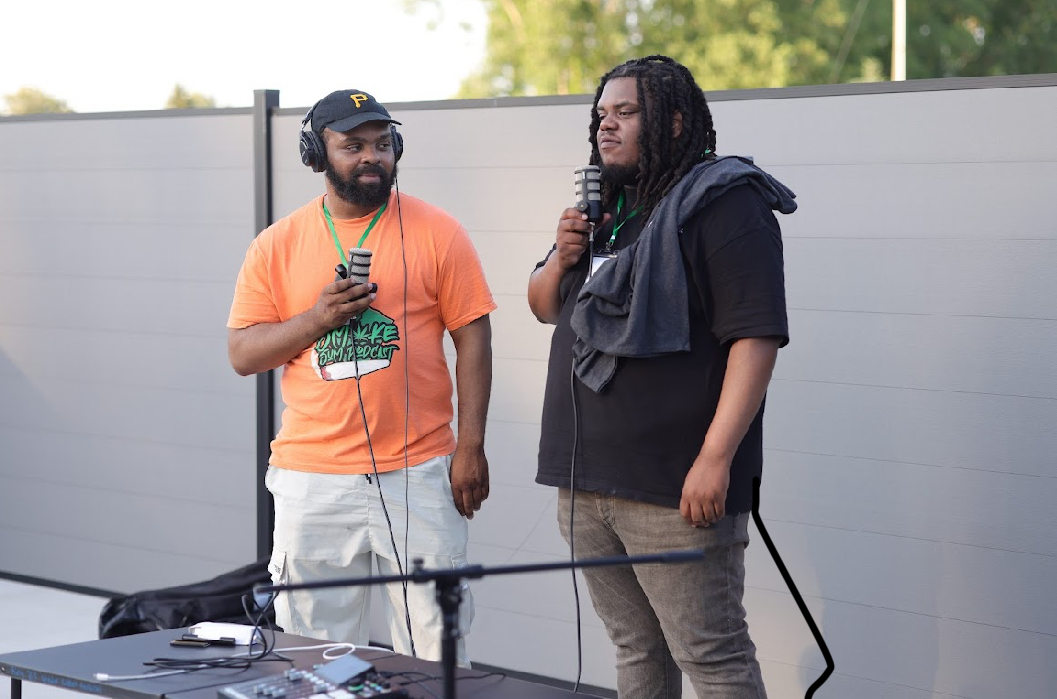 Two men with microphones stand outdoors near a gray fence, one wearing an orange shirt and the other in black, talking.