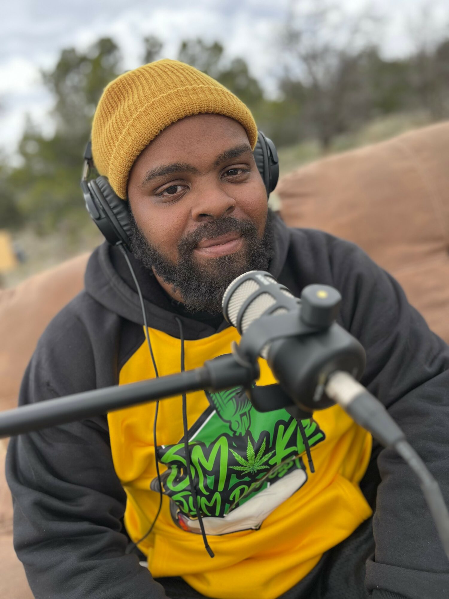 Man with a beard wearing a yellow beanie and headphones, smiling outdoors with a microphone in front.