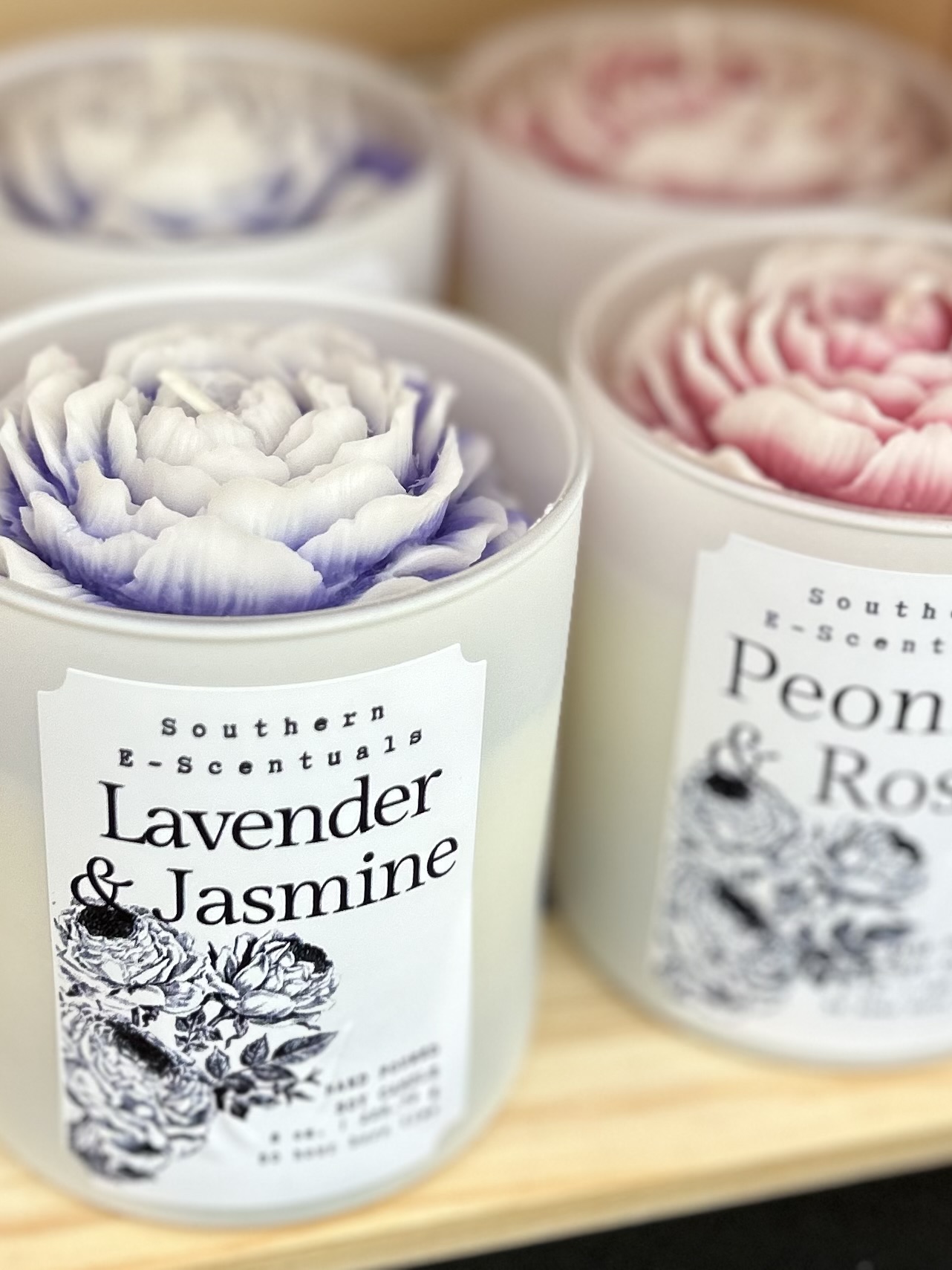 Four candles with flower-shaped wax, lavender, jasmine, peony, and rose scents, in white containers on a wooden surface.