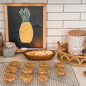 Baked cookies on a wire rack, with a framed pineapple drawing, bowls, and a jar on a kitchen counter.