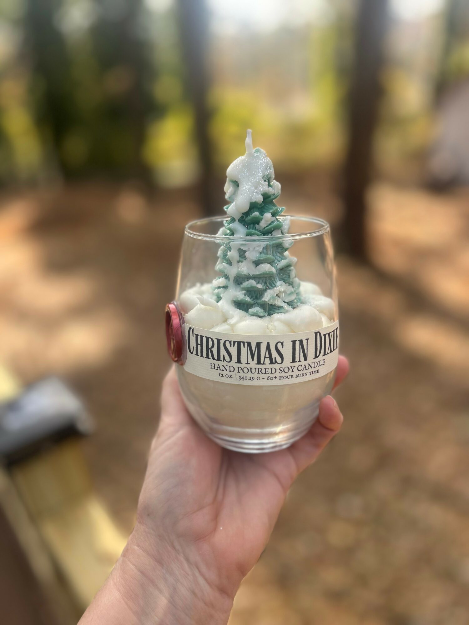 Hand holding a glass with a small Christmas tree-shaped candle inside outdoors, blurred background of trees and sunlight.