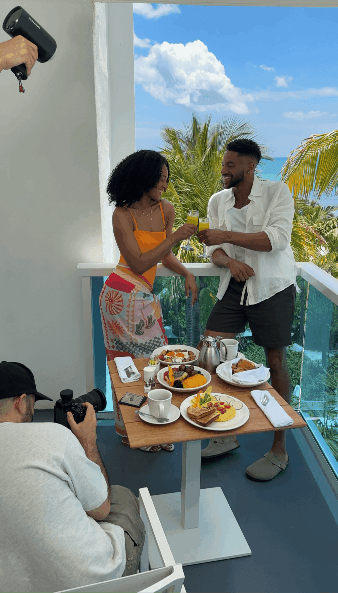Two people exchanging a drink on a balcony with a table of food, a person taking photos, and a blue sky with clouds.