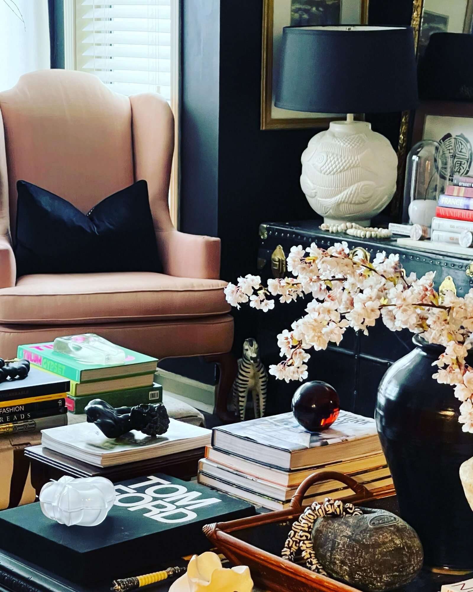 Living room with pink armchair, black pillow, side table, lamp, and decorative items, including a large black vase with flowers.