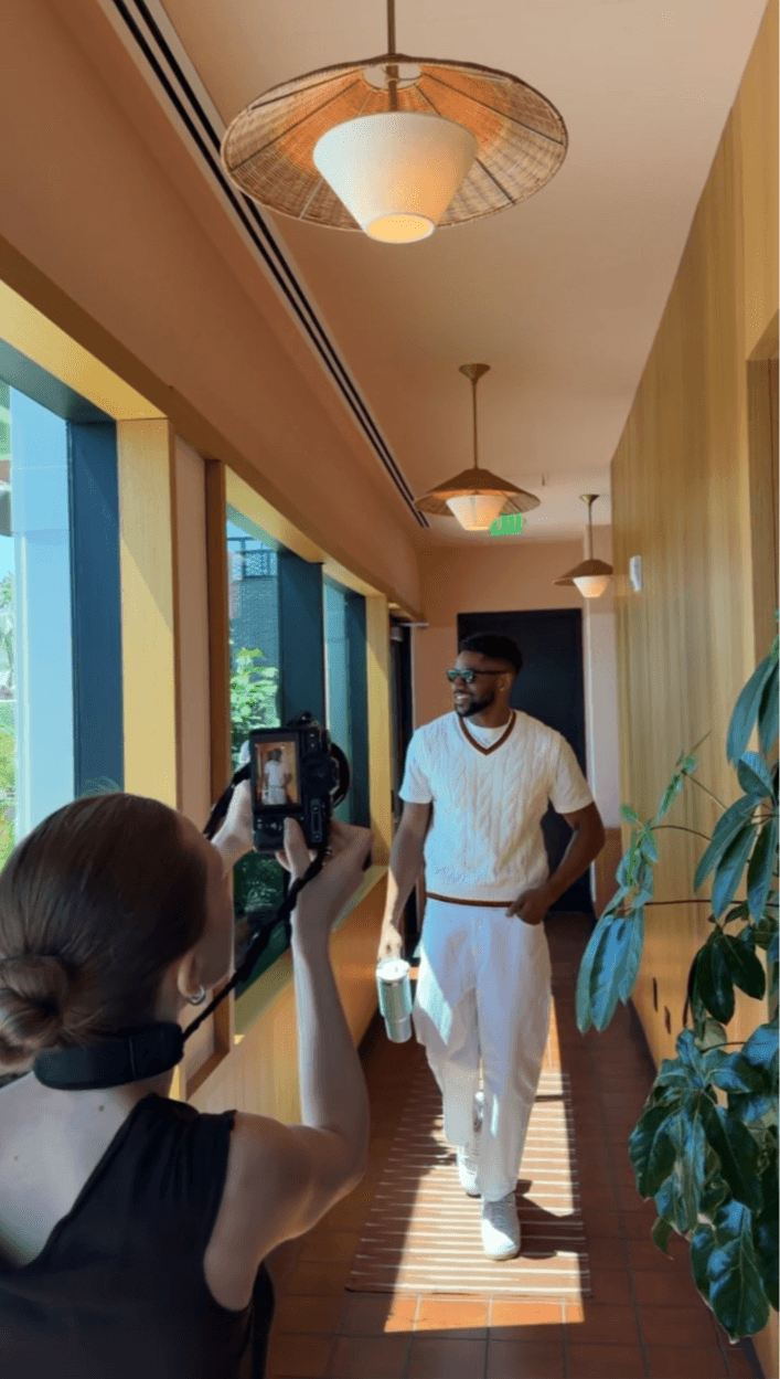 Person taking a photo of a man walking in a brightly lit corridor with large windows and hanging ceiling lights.