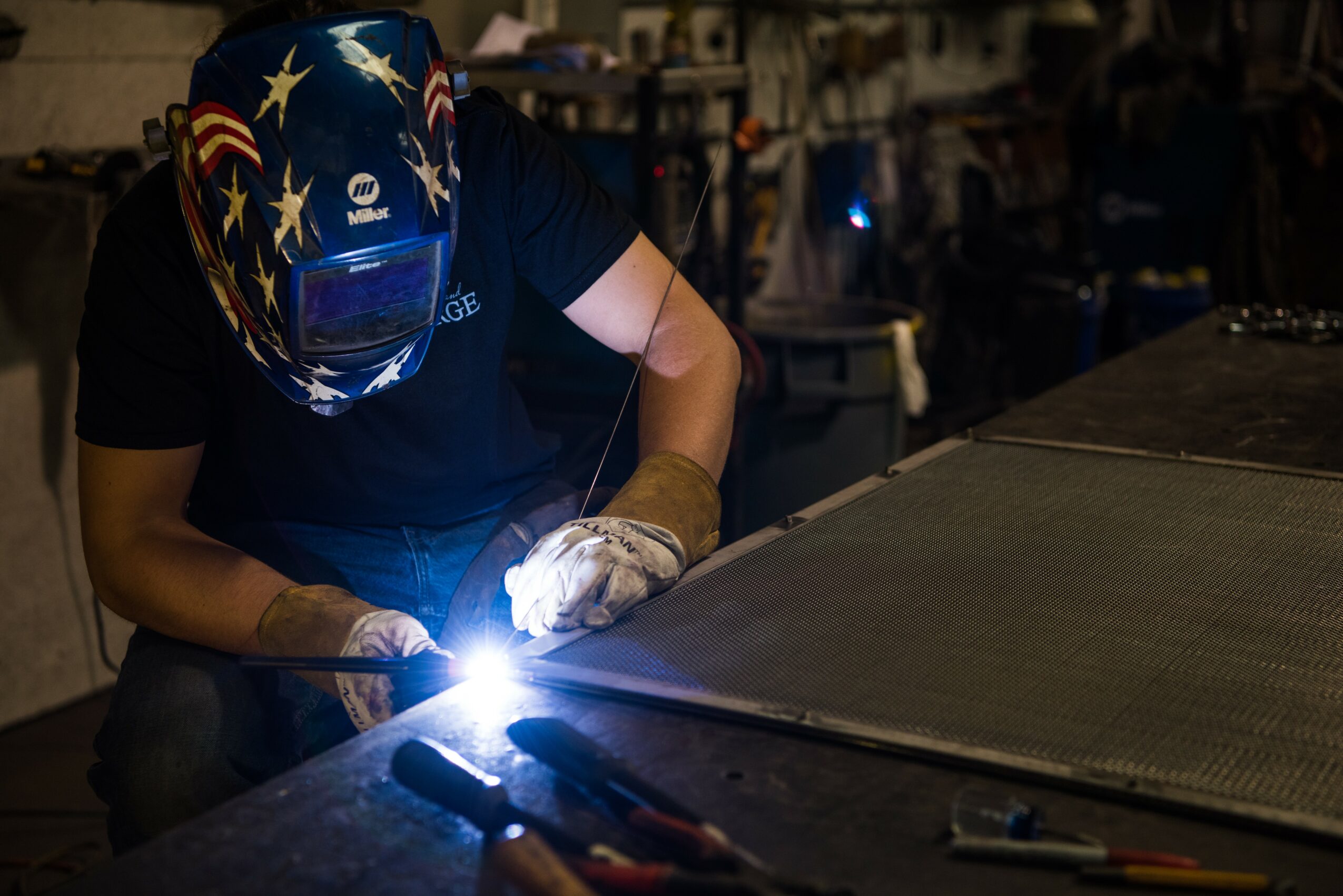 Person welding metal, wearing a helmet with stars and stripes, working in a workshop with tools nearby.