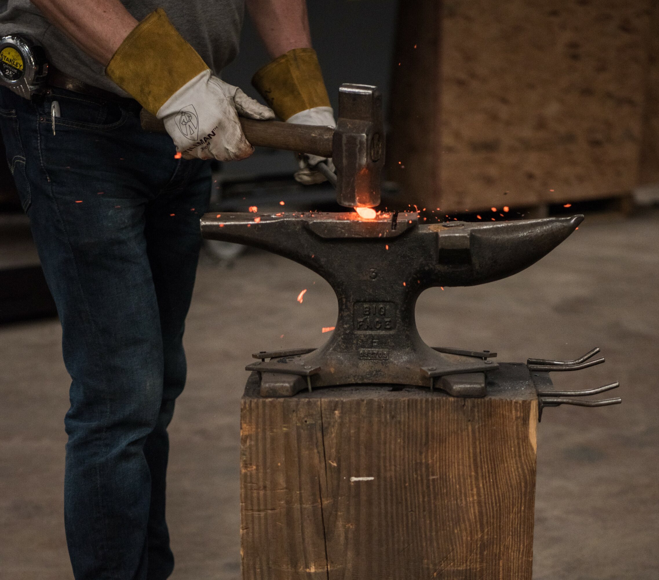 Person wearing gloves hammering metal on an anvil with sparks flying, standing next to a wooden block.