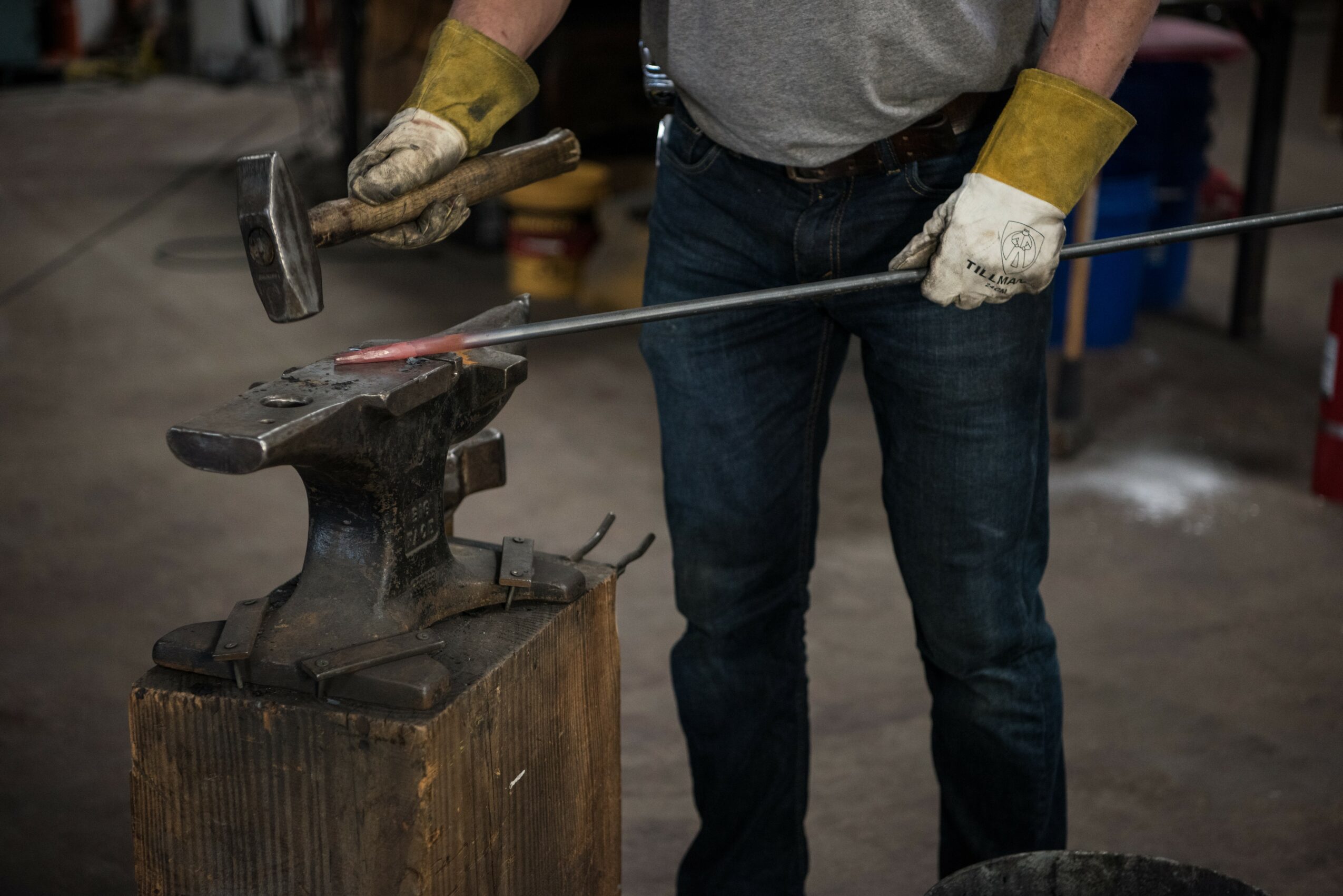 Person wearing gloves hammering a metal rod on an anvil in a workshop.