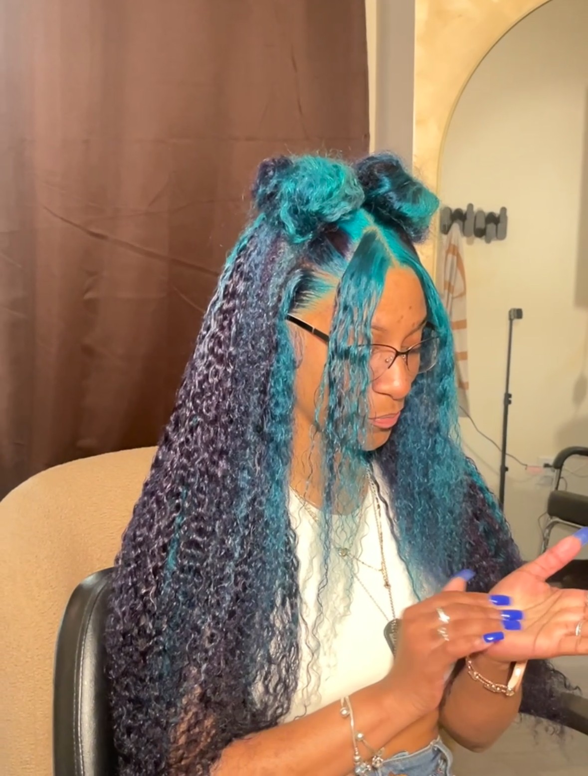 Woman with long, curly blue and purple hair, wearing glasses, looking at her phone, sitting indoors.