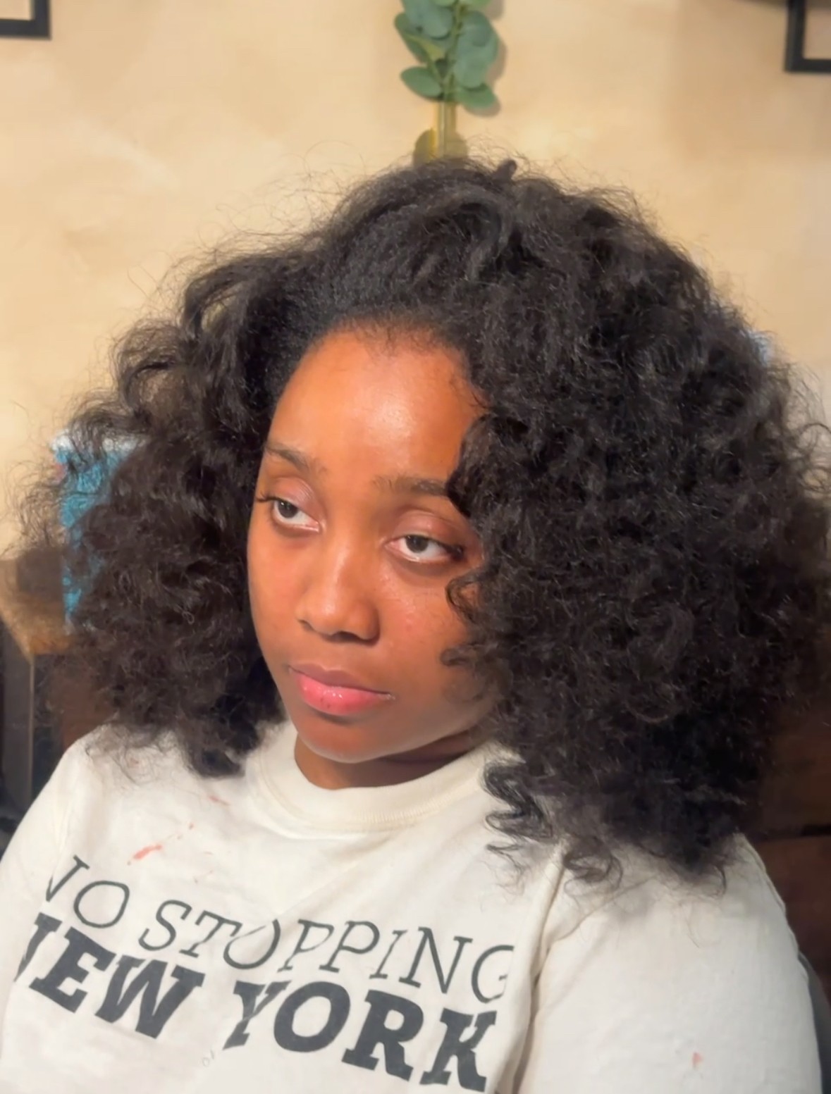 Young woman with voluminous curly hair looking to the side, wearing a white shirt with text, indoors.