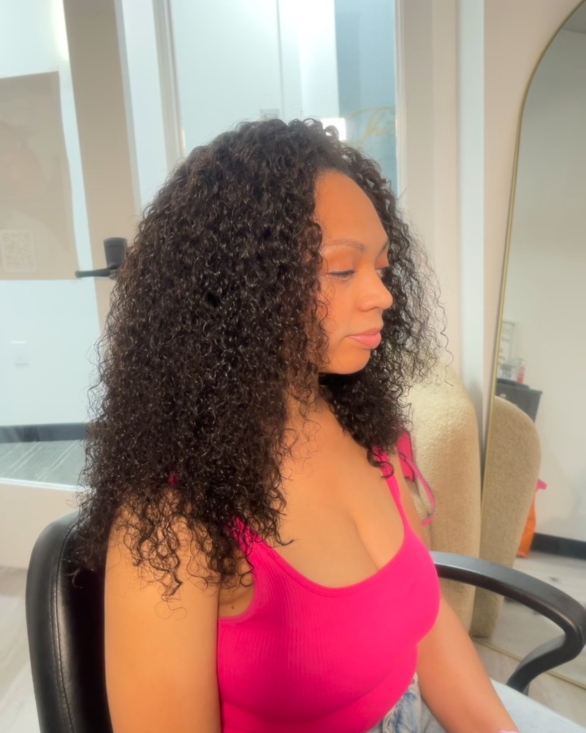 Side view of a woman with curly hair wearing a pink top, sitting in a salon chair, looking to the side.