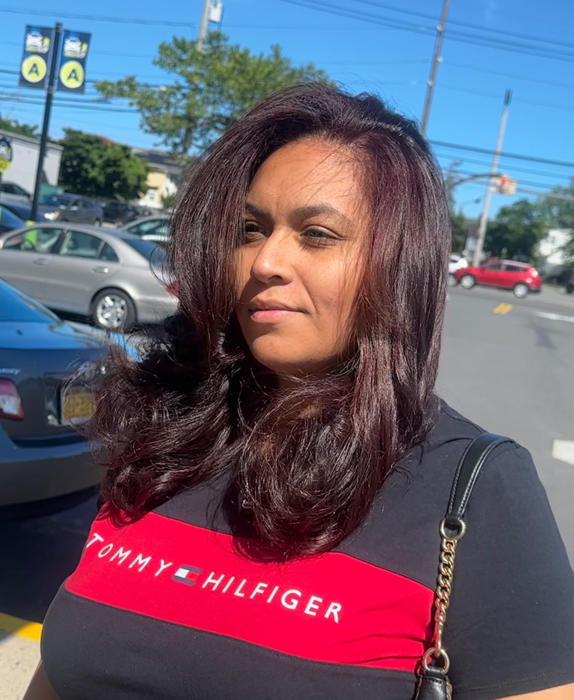 Woman with long dark hair standing outdoors in a parking lot, wearing a black and red Tommy Hilfiger shirt.