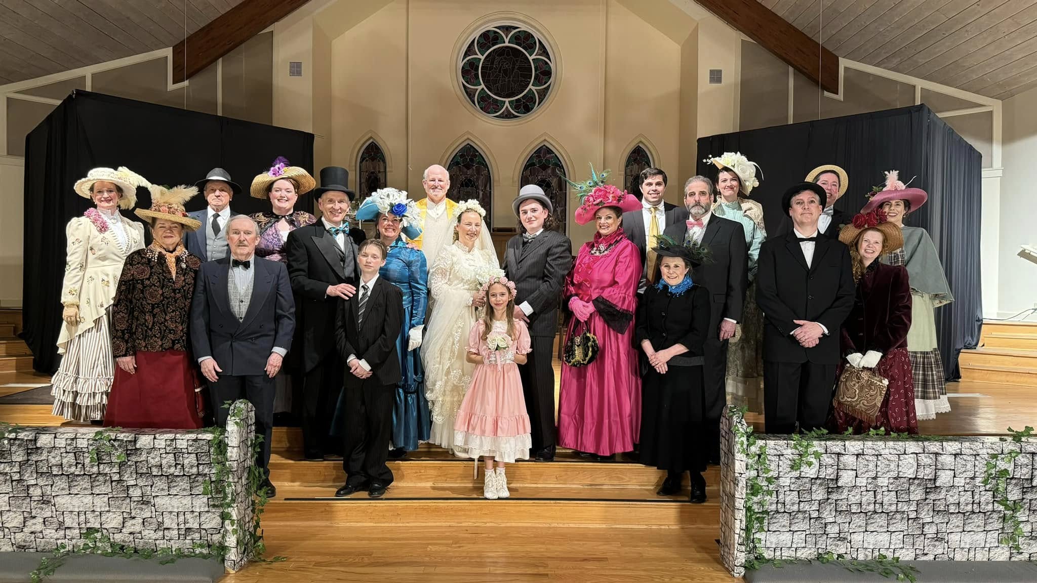 Group of people dressed in vintage clothing on stage, with a church interior background, including a circular window.