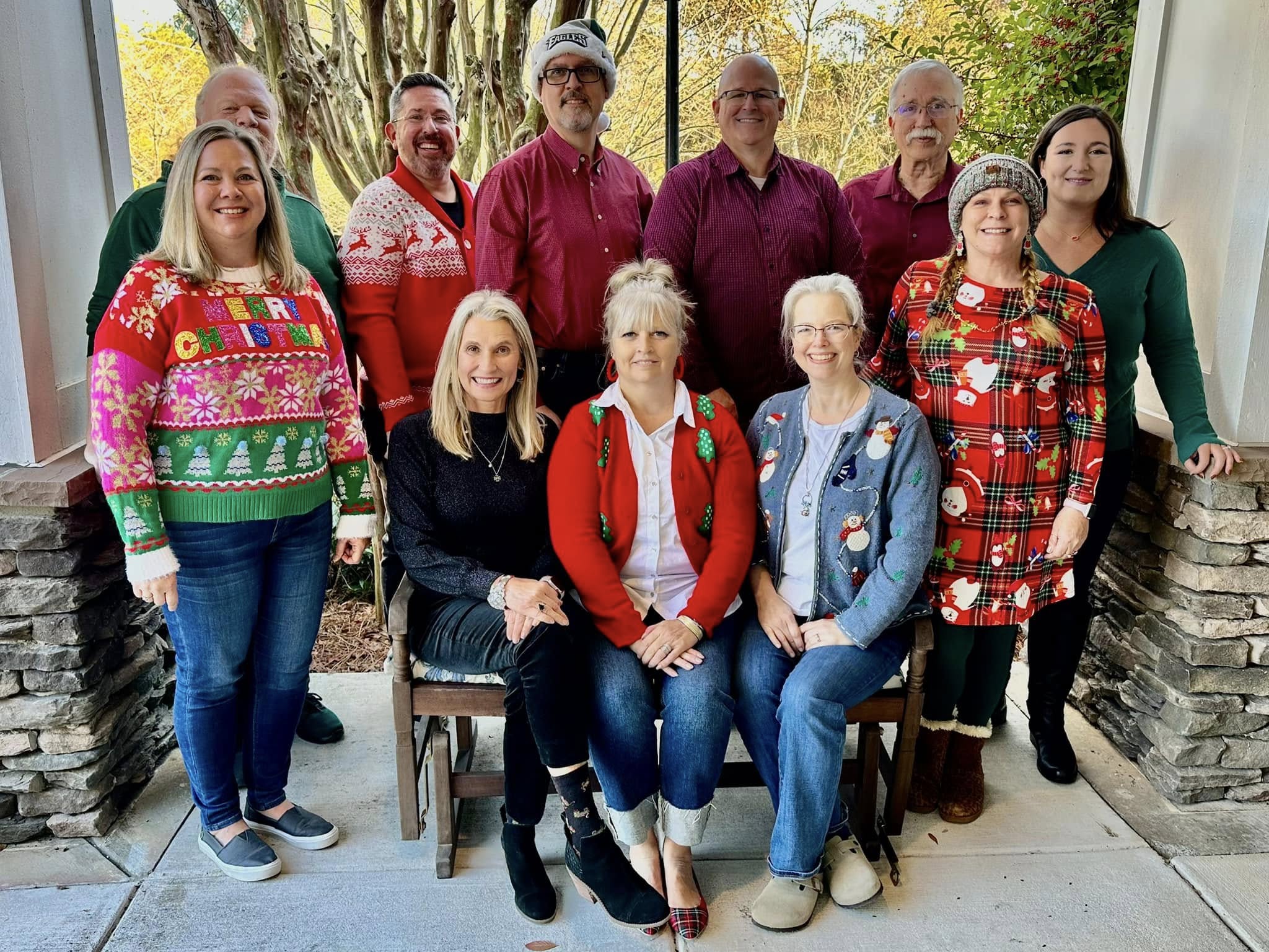 Group of ten people, some wearing festive sweaters, standing and sitting outdoors with trees in the background.