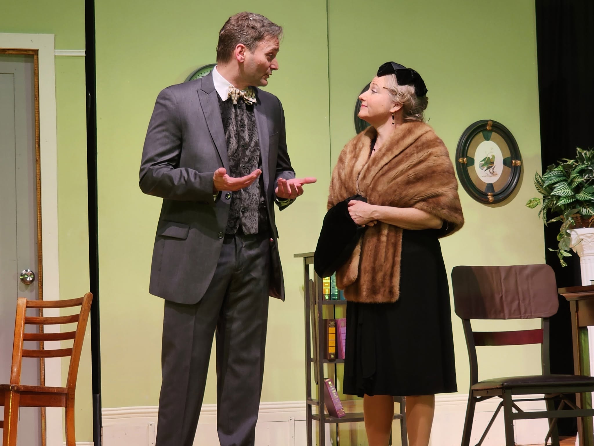 Two people, a man and a woman, stand and converse in a room with green walls, chairs, and a plant.