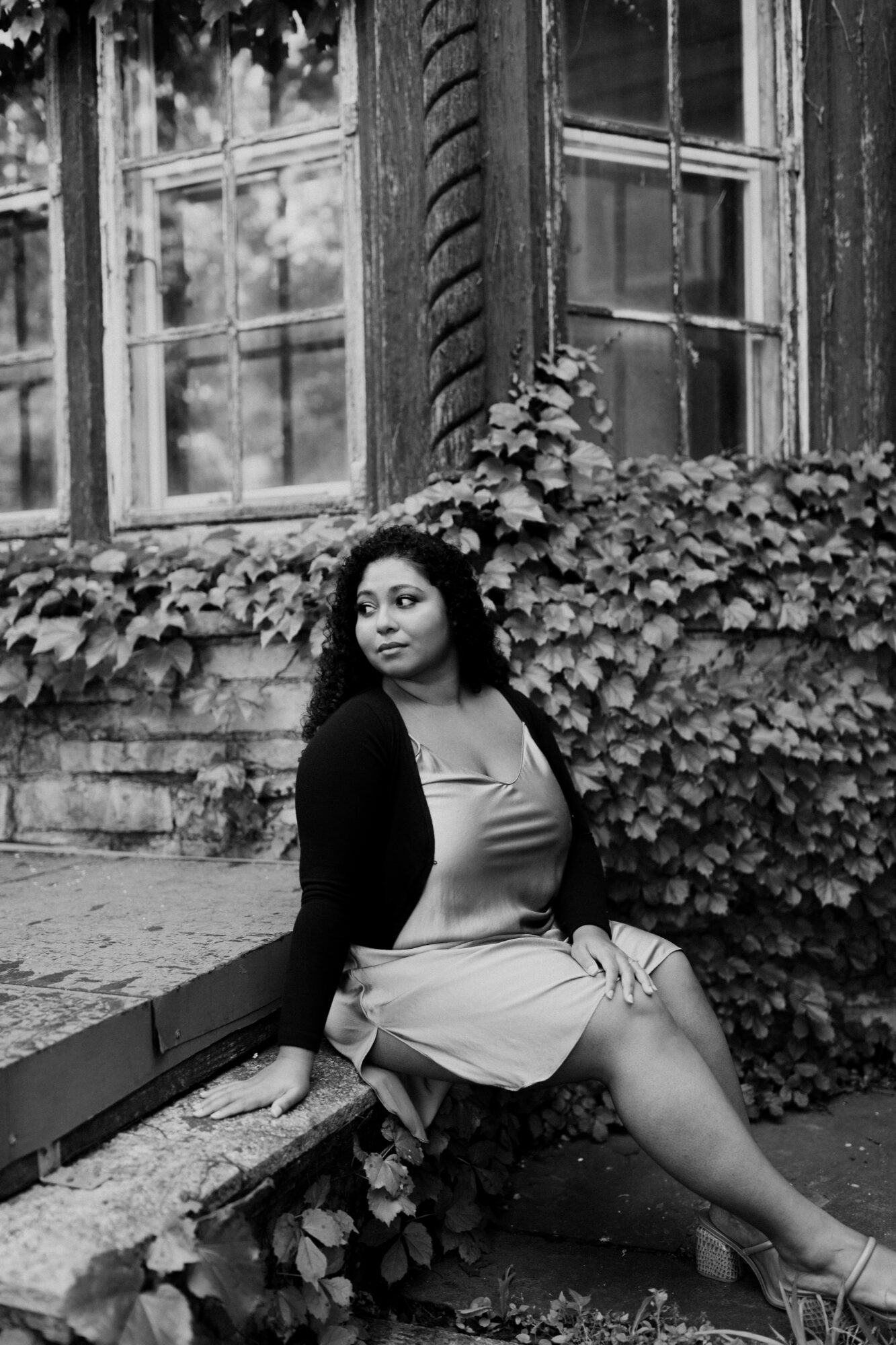 Woman sitting on a stone ledge in front of a building with ivy-covered walls and large windows.