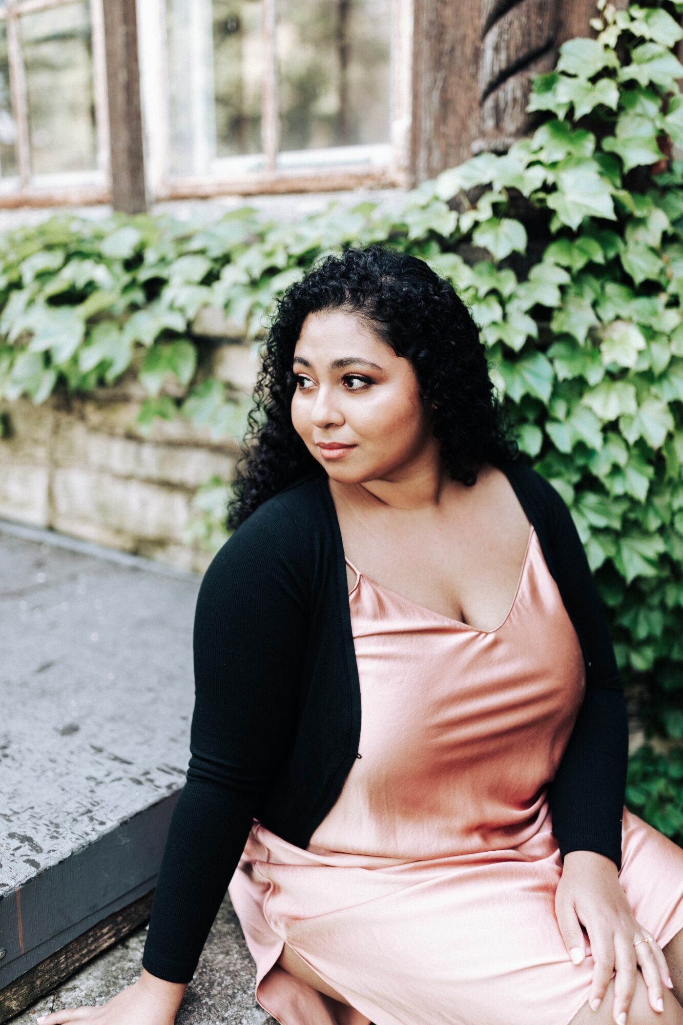 Woman with curly hair sitting outdoors near a brick wall covered in ivy, wearing a pink dress and black cardigan.