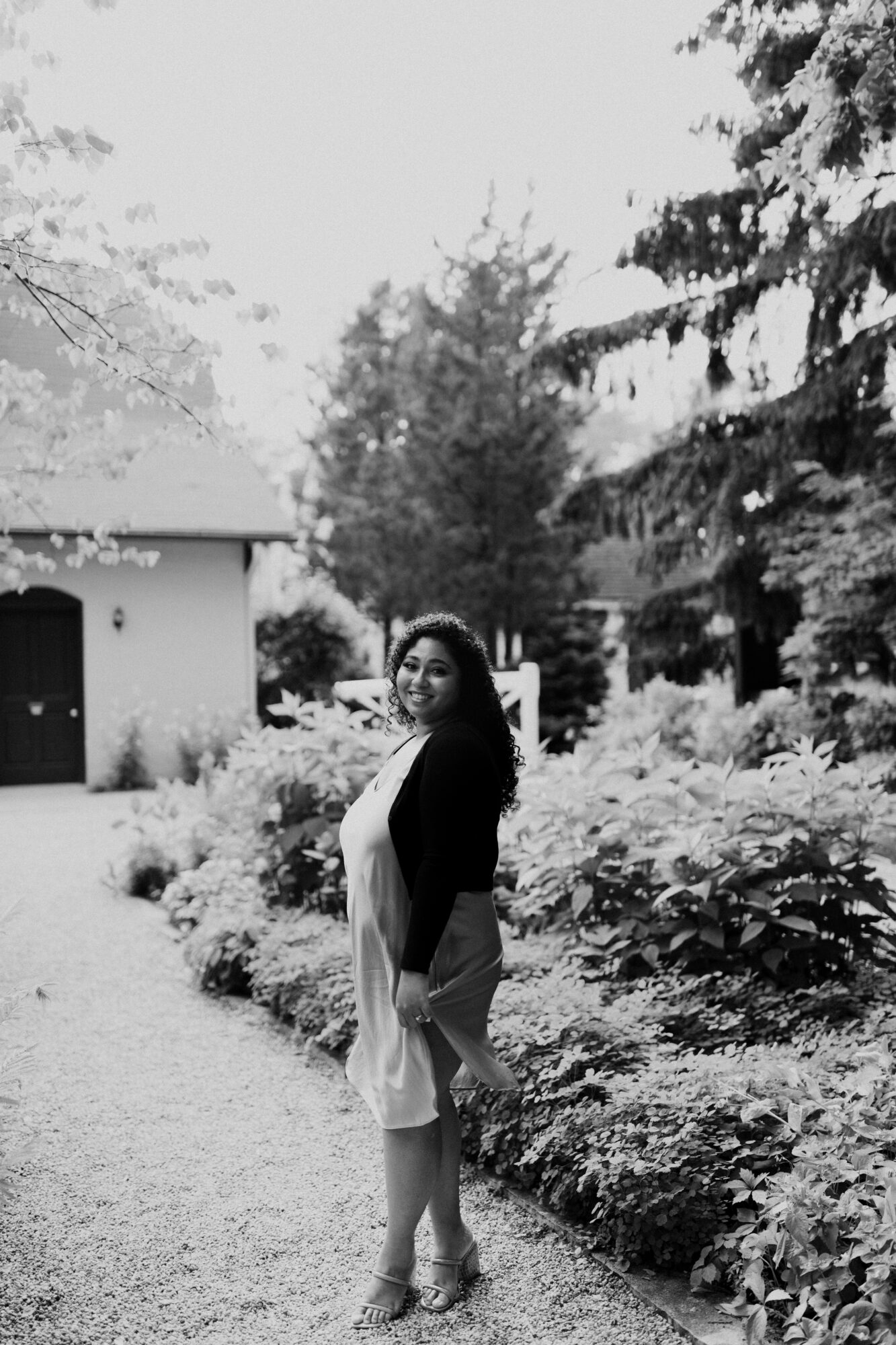 Woman standing outdoors on a pathway surrounded by trees and bushes, smiling, in black and white.