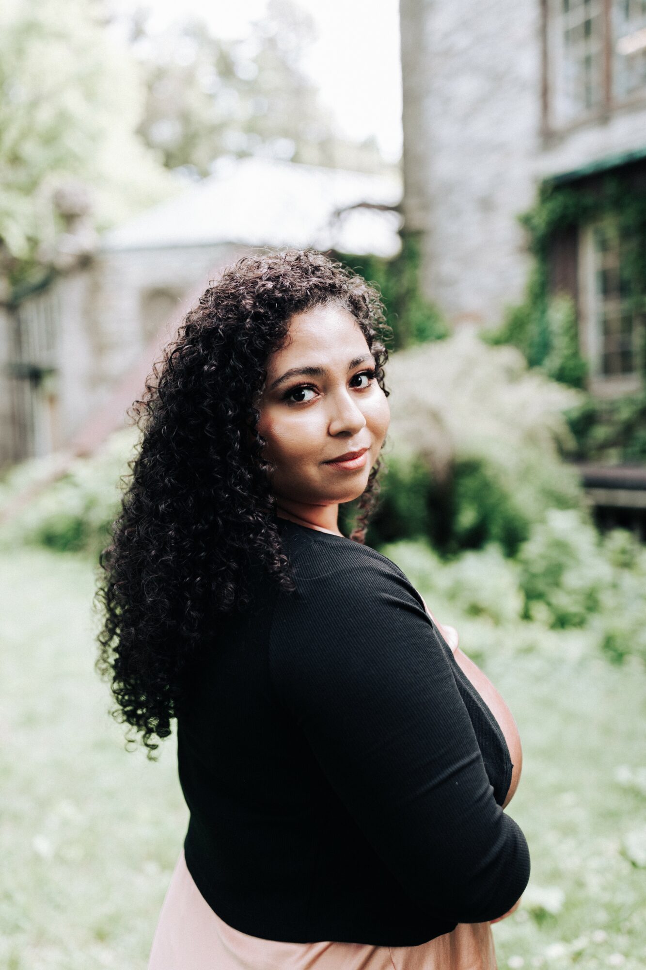 Woman with curly hair standing outdoors in a garden, looking over her shoulder, with greenery and a building in the background.