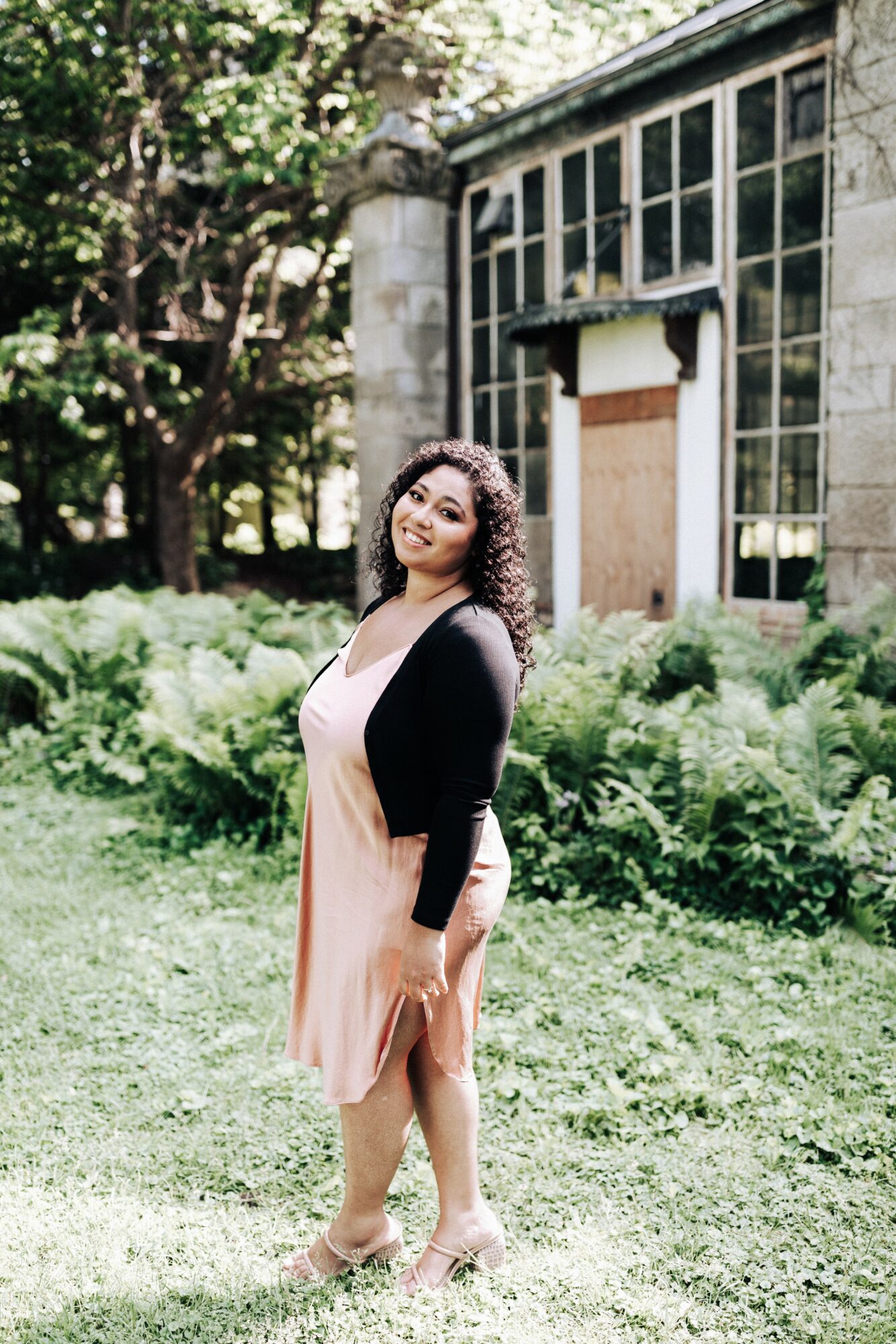 Woman with curly hair standing outdoors in front of a building with large windows, surrounded by greenery.