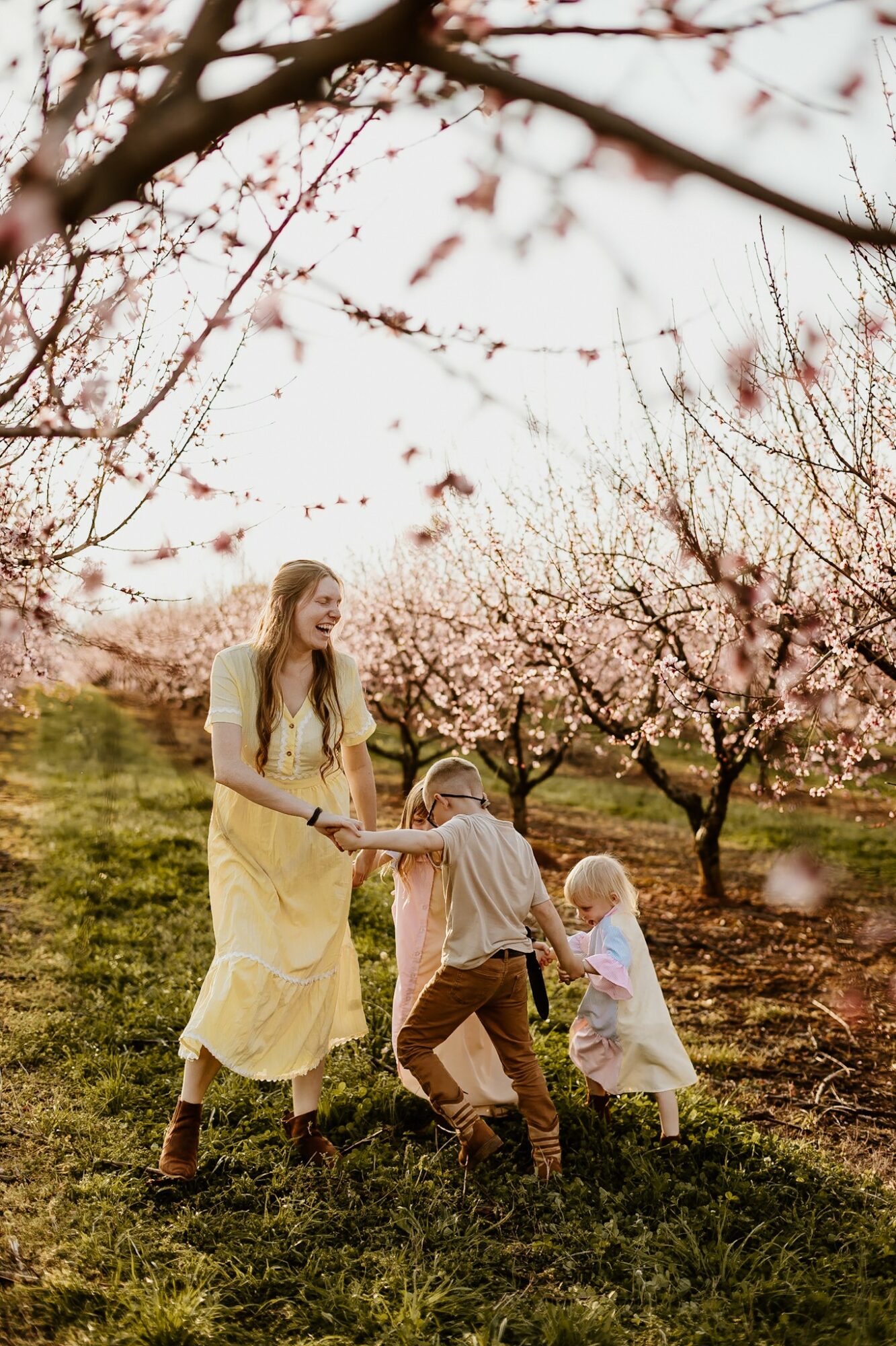 A woman and two children playing in a blooming orchard with pink flowers, green grass, and trees in the background.