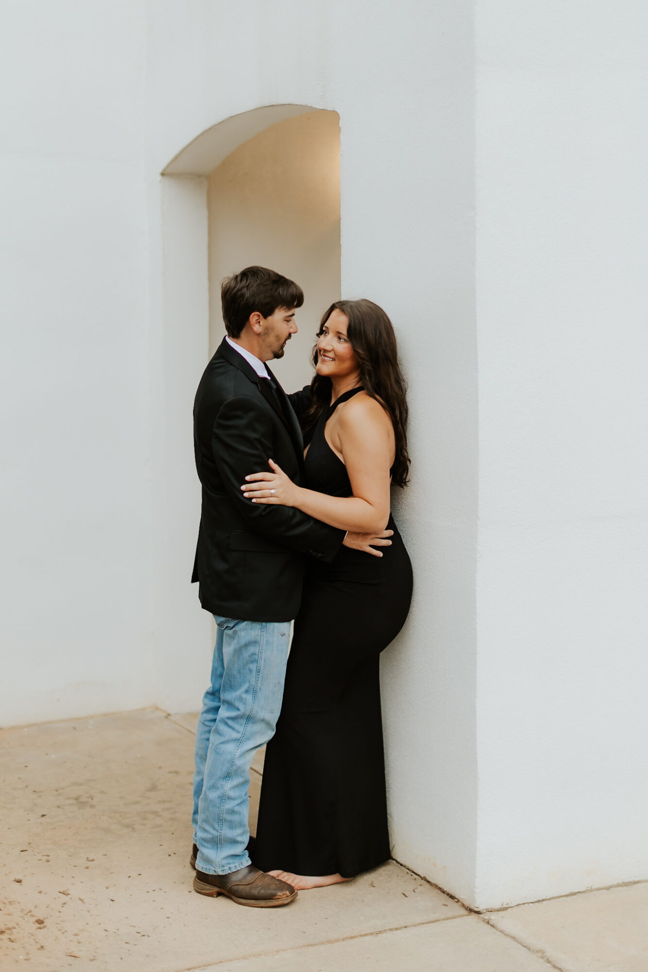 A man and woman stand close, smiling and looking at each other, in a minimalistic white space with an arched opening.