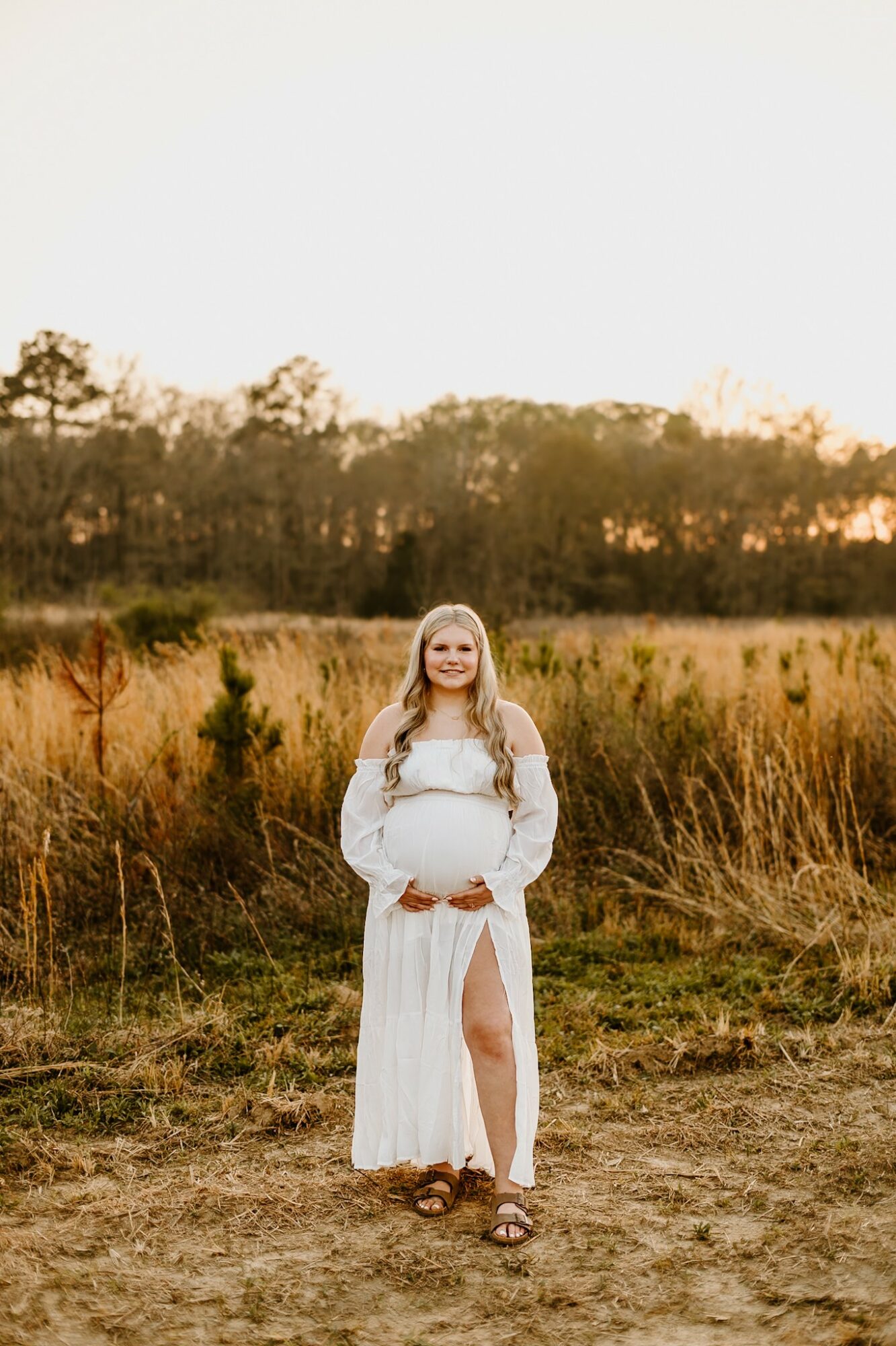 Pregnant woman in a white dress standing outdoors in a field with trees in the background, smiling, during sunset.